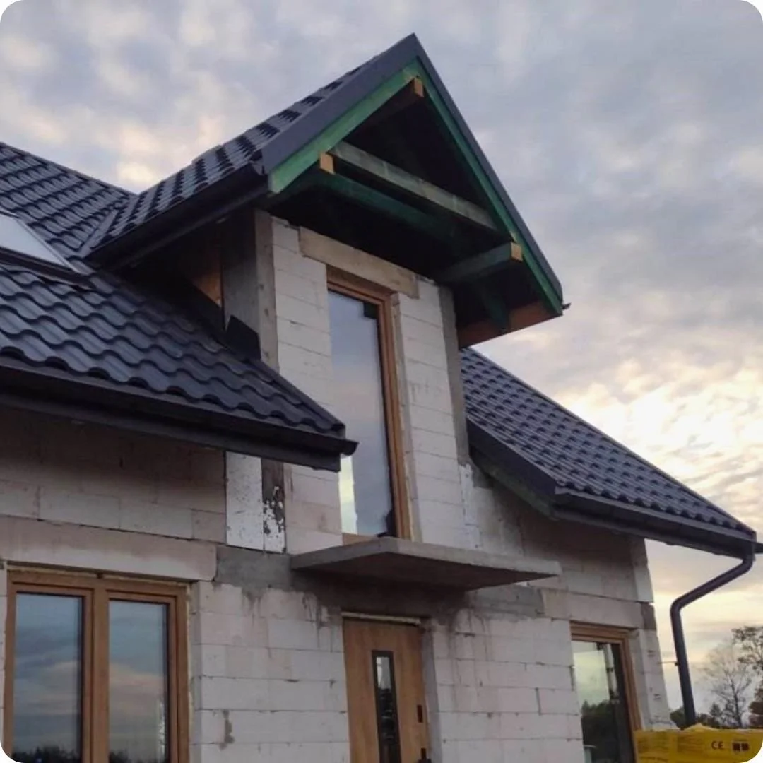 Dach z czarnej blachodachówki z wykuszem i oknem balkonowym w stanie surowym.