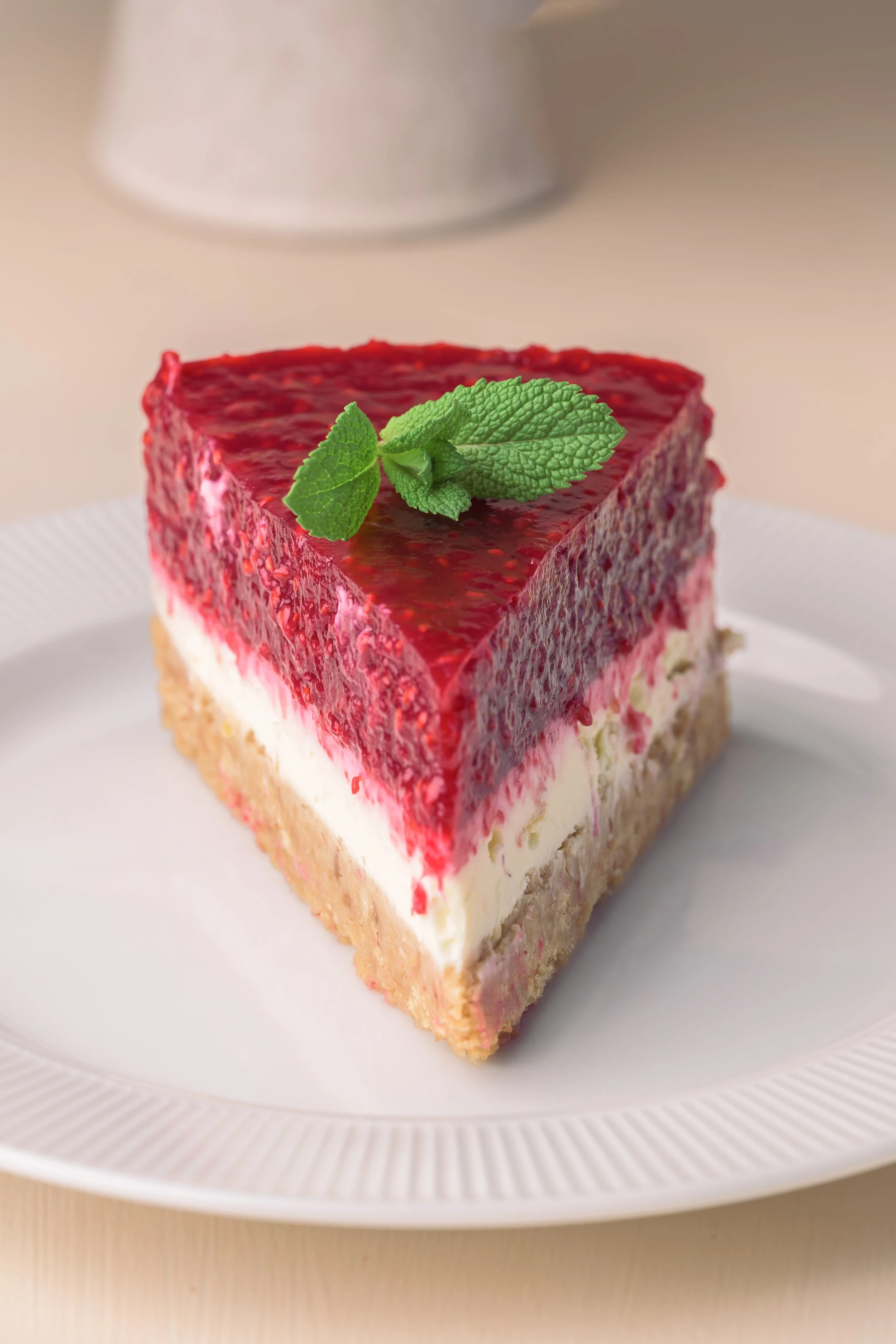 Ein Stück No-Bake Himbeer Cheesecake mit einer Schicht aus roten Beeren, einer weißen Creme-Schicht und einem crustartigen Boden, garniert mit Minzblättern auf einem weißen Teller.