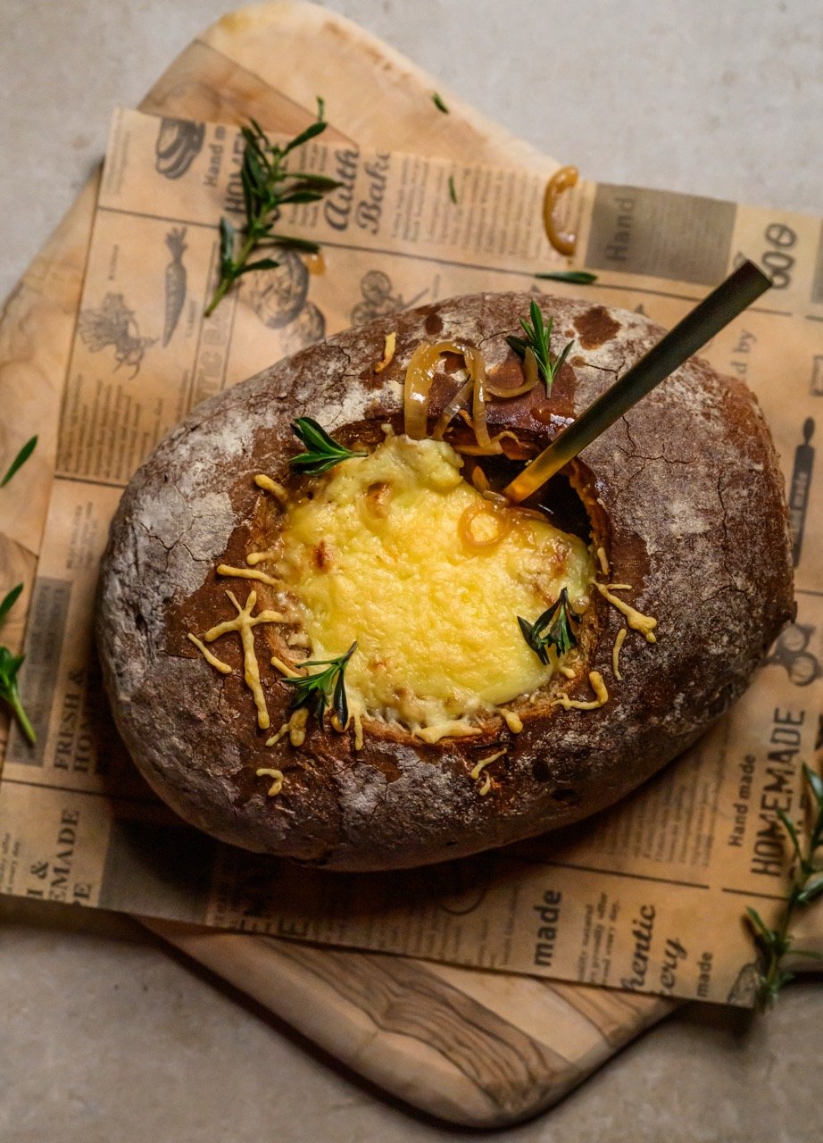Ein ausgehöhltes rustikales Brotgefüllte französische Zwiebelsuppe Gericht getoppt mit Käse und Kräutern serviert auf Zeitungspapier.