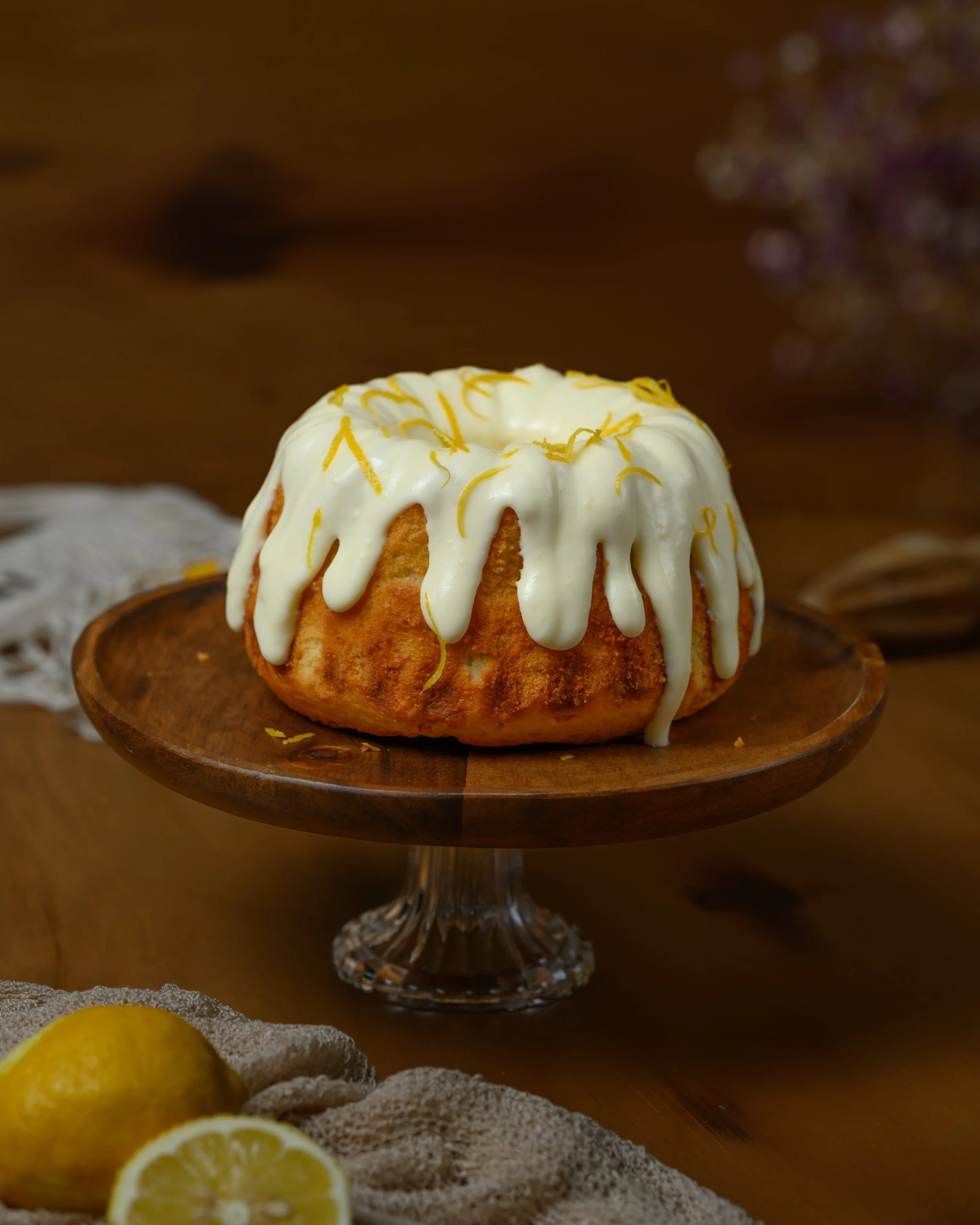 Ein Zitronenkuchen mit weißer Glasur auf einem Holzsockel, Dekor mit Zitronenschale, auf einem Holztisch mit Zitronen und Stoff.