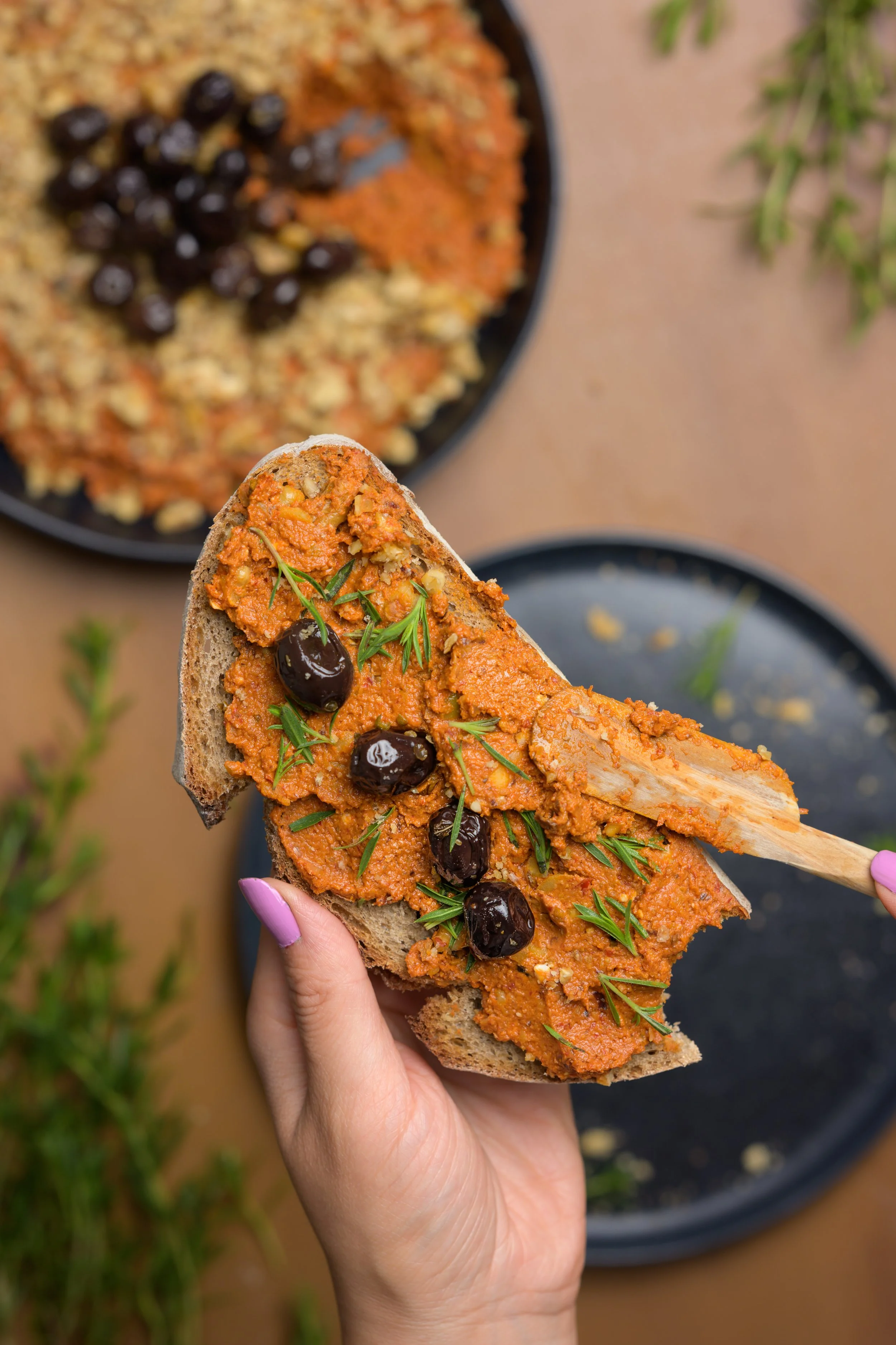 Hand, die eine Scheibe Brot mit veganer Oliven-, Paprika- und Tomatenpaste, schwarzen Oliven und Thymian hält, im Hintergrund eine Schüssel mit der gleichen Paste