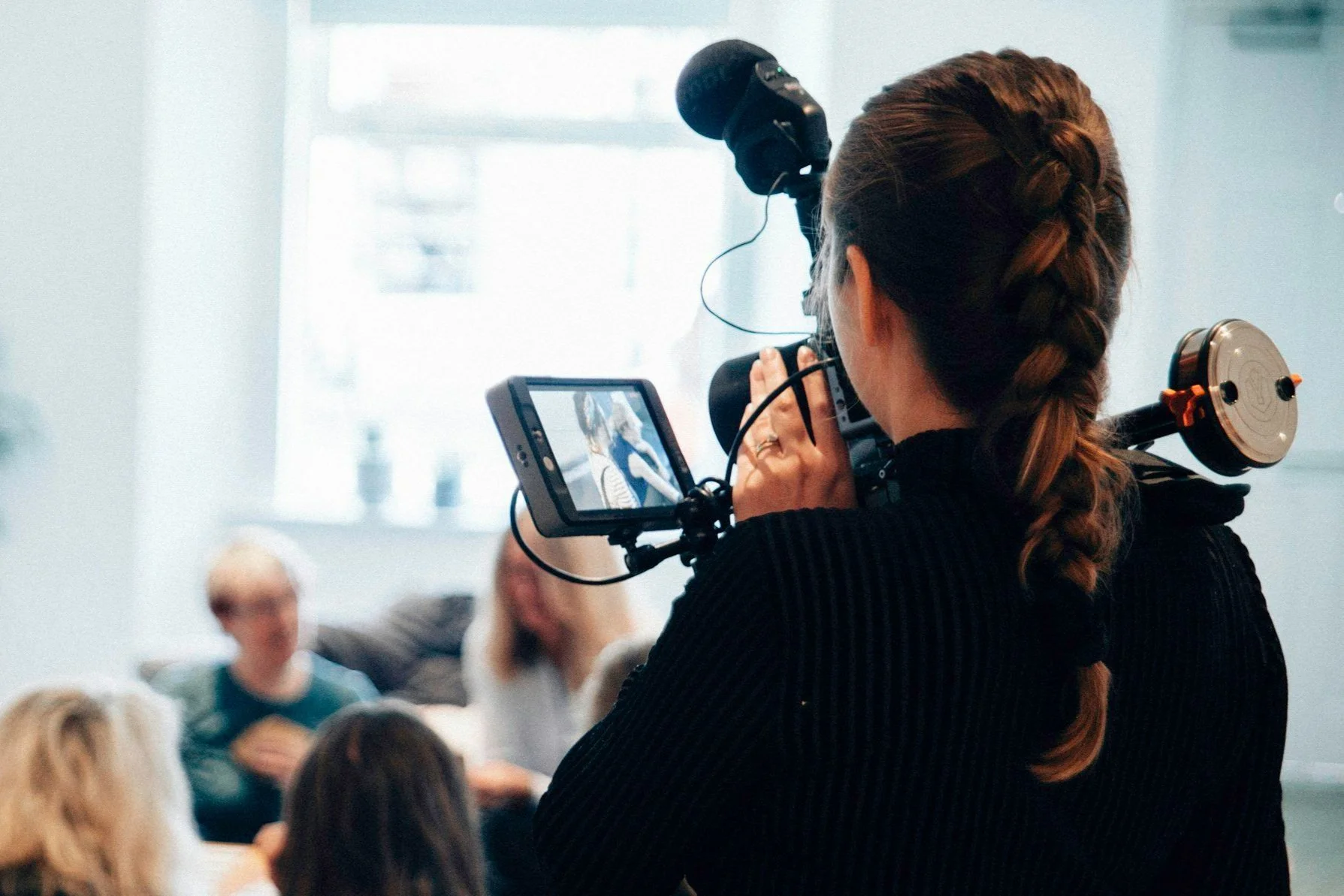 A woman with braided hair filming a discussion with a camera in a bright room, with three older women seated and talking in the background.