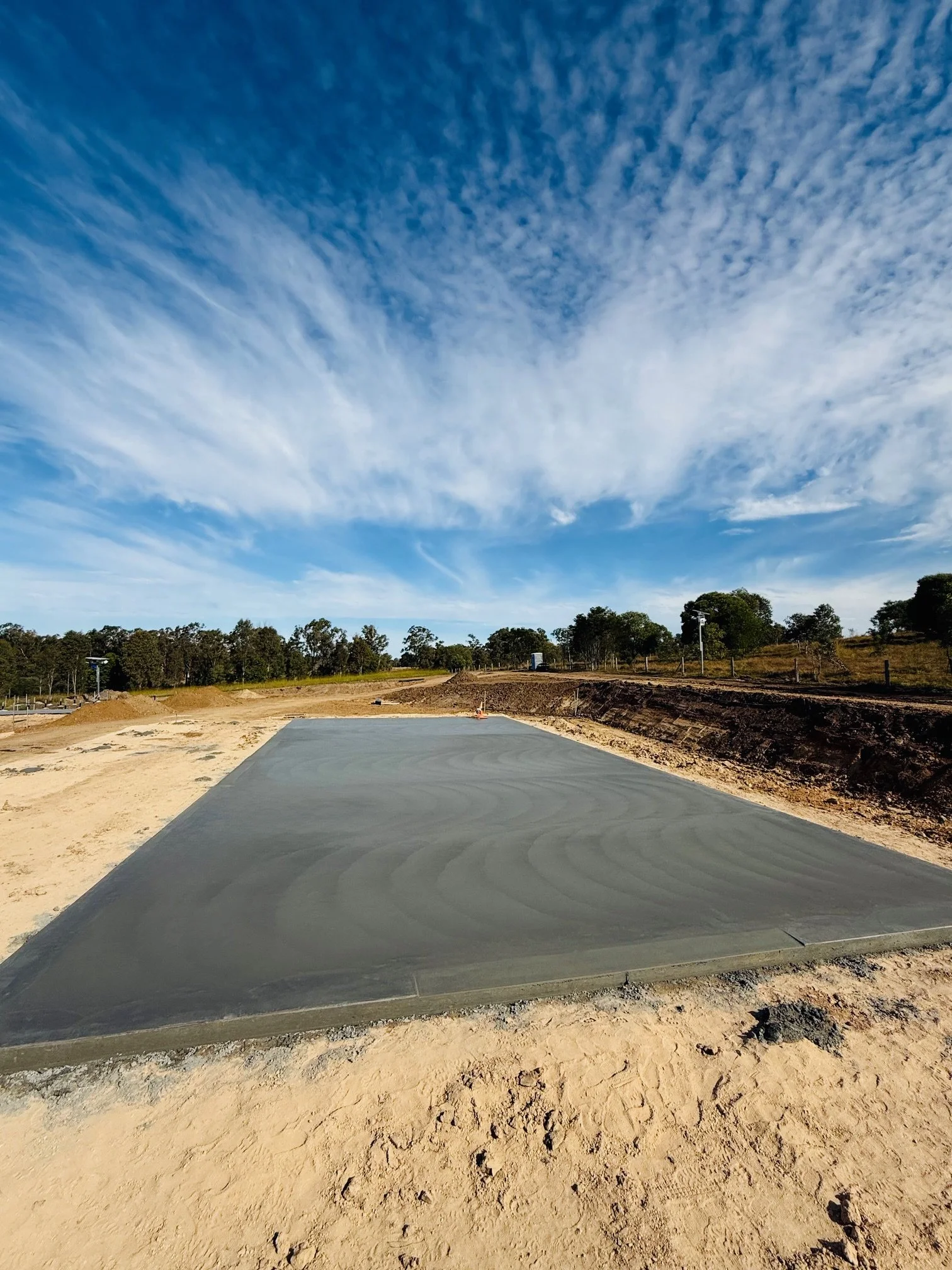 A freshly poured concrete slab on a construction site with trees and a blue sky with clouds in the background.