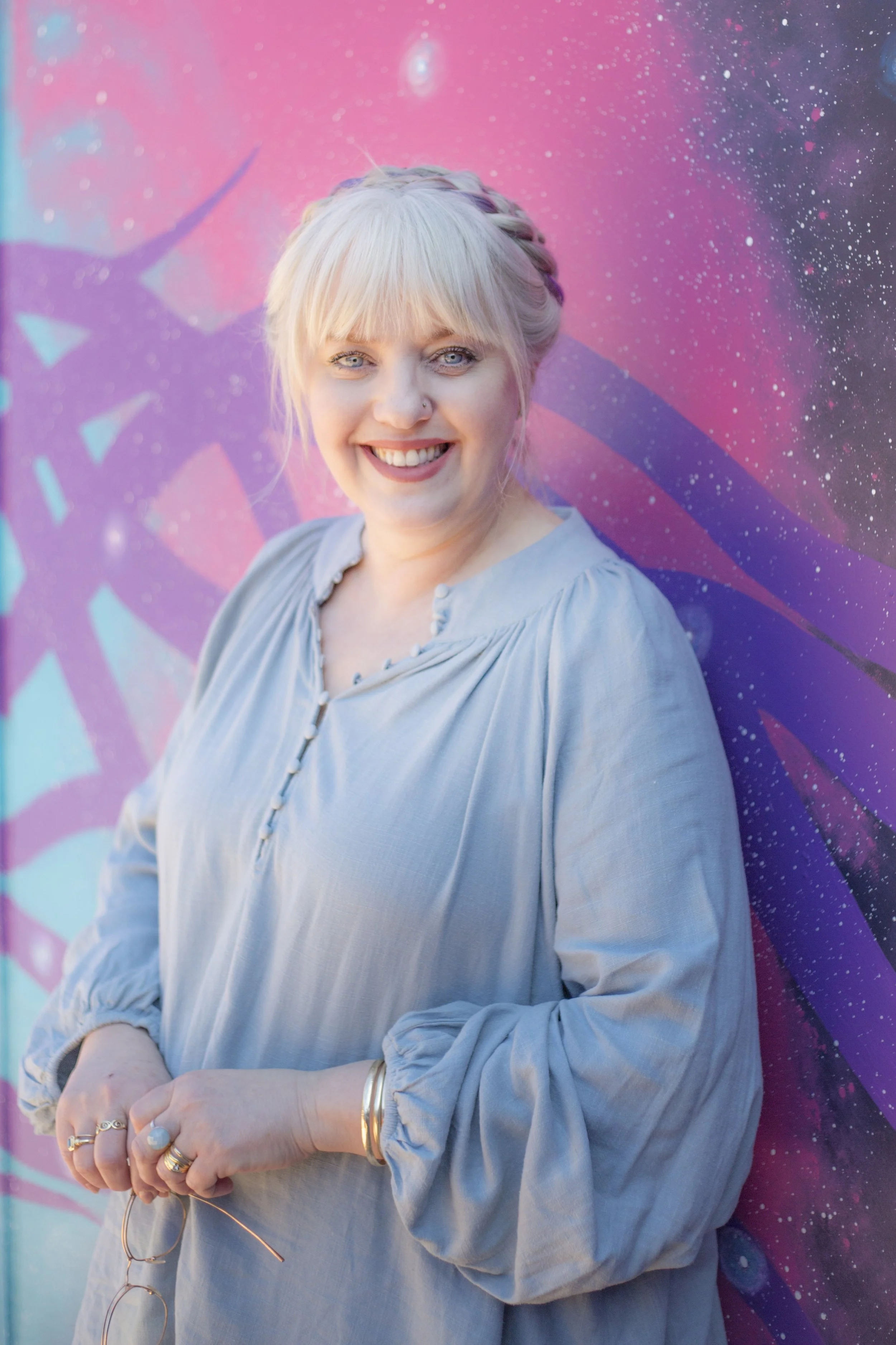 White woman with blonde hair and blue eyes smiles warmly at the camera, she is leaning back against a wall with a bright galactic theme.