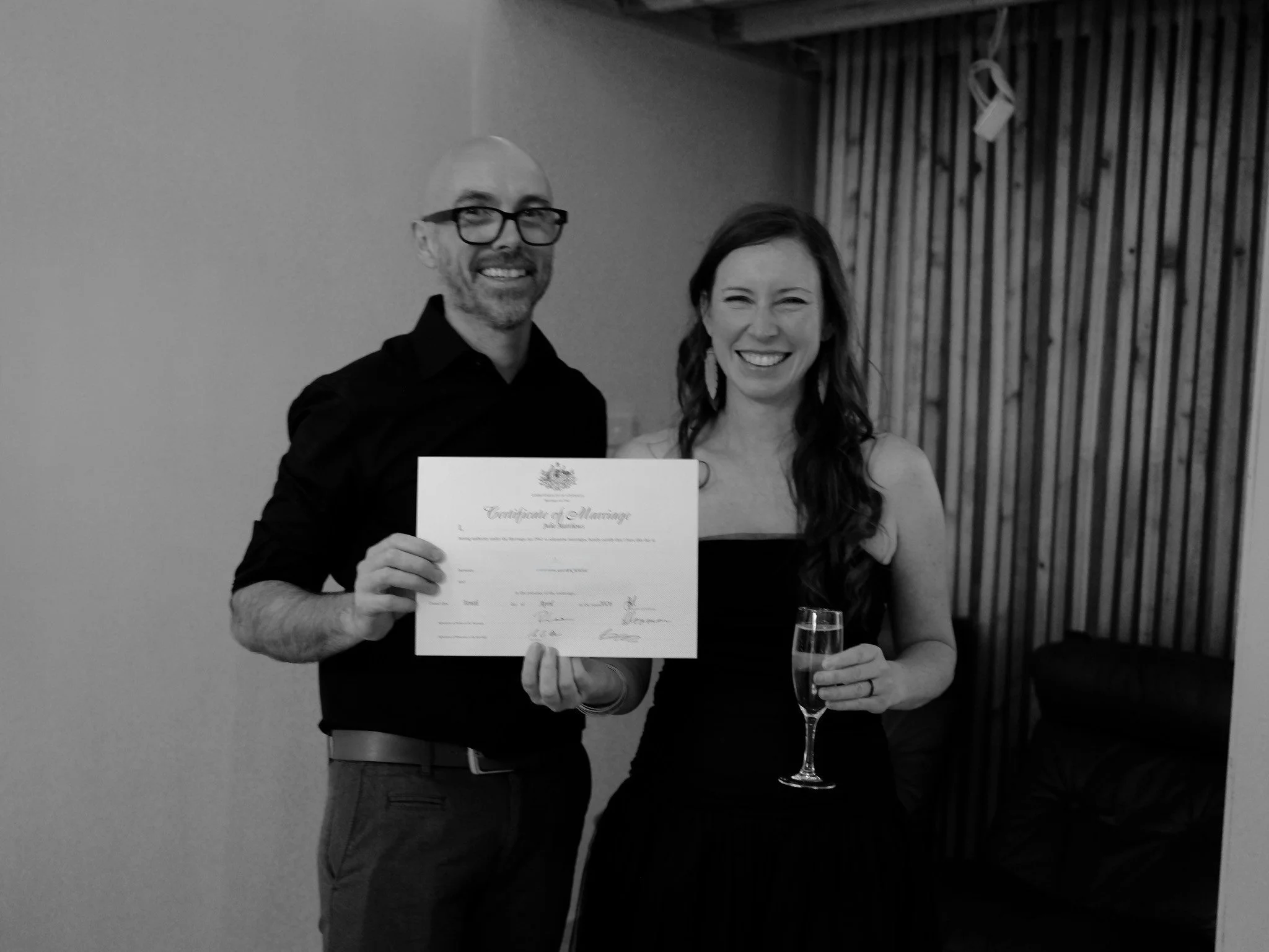 Couple stand together holding wedding certificate with huge relaxed smiles. Bride is holding a glass of bubbly wine