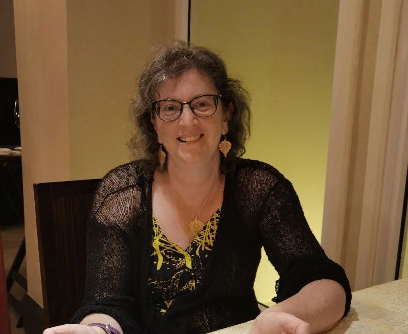 A smiling woman with curly gray hair, glasses, and wearing a black and yellow outfit, sitting at a dining table indoors.