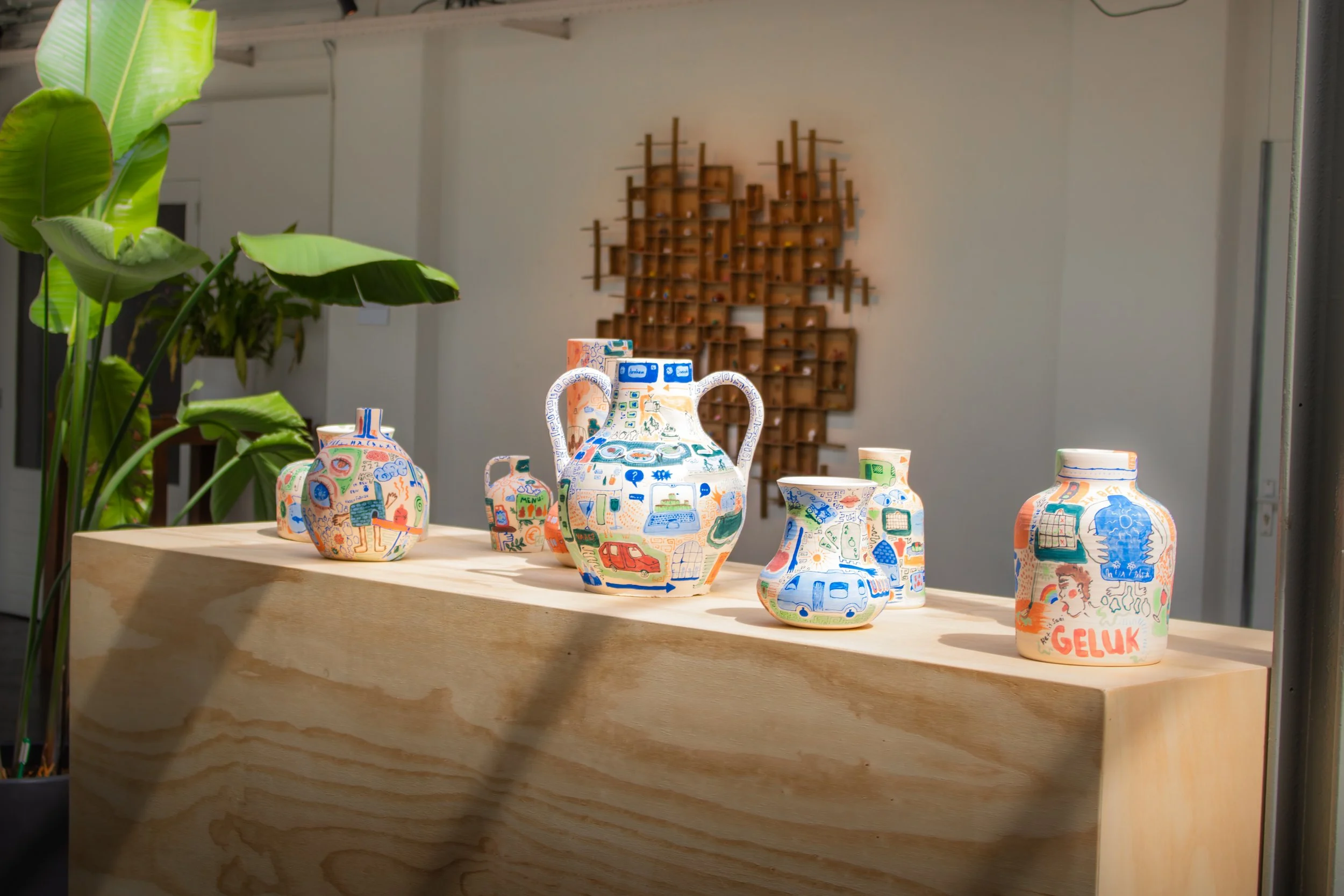 Kleurrijke keramieken vazen en potten met tekeningen op een houten tafel, in een binnenruimte met grote groene planten en een houten wandsculptuur op de achtergrond.