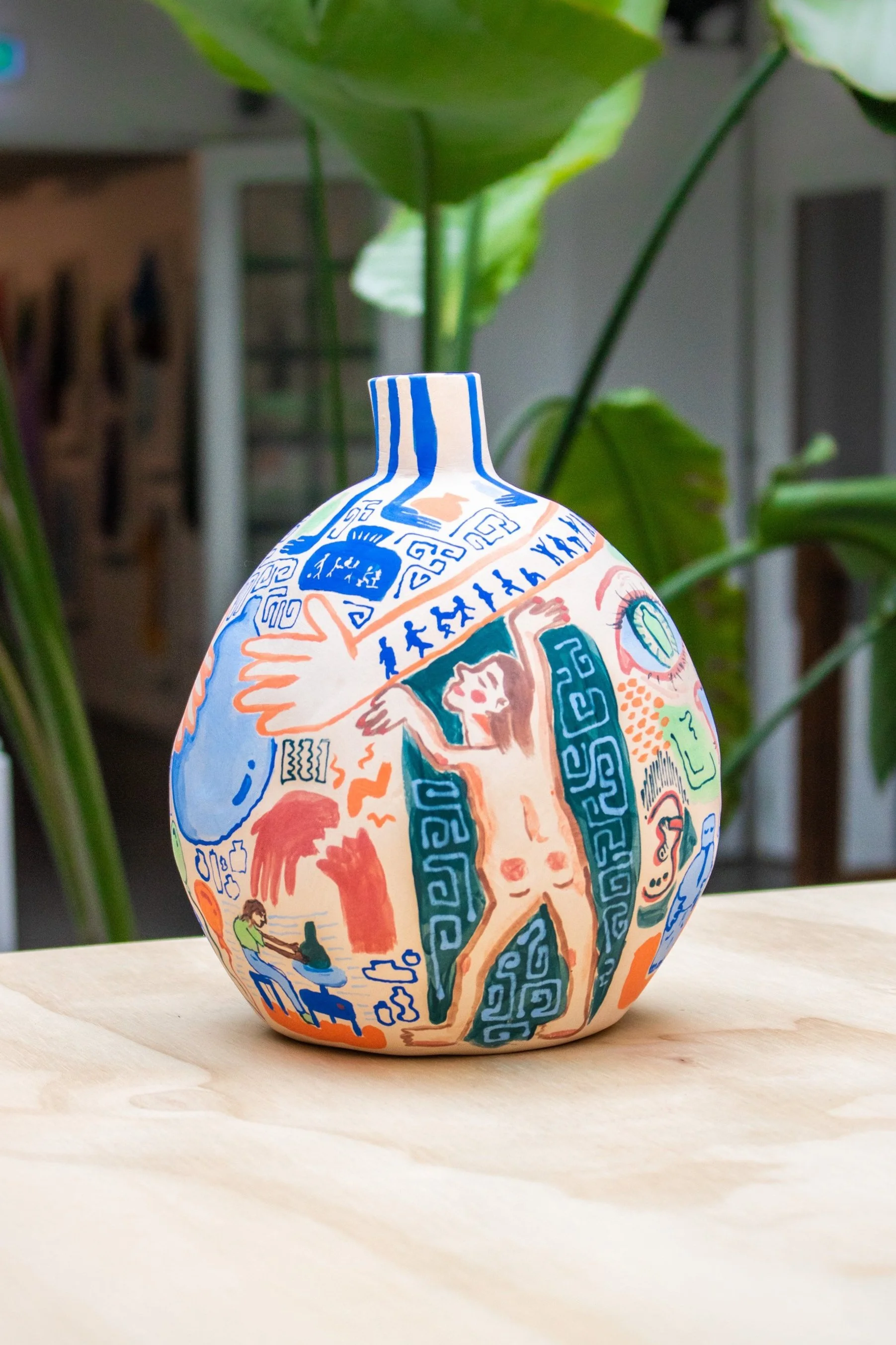 Sieradeker aangoekleed met kleurrijke, abstracte en figuratieve tekeningen, waaronder een slapende mens, gezichten, en symbolen, op een houten tafel met groene planten op de achtergrond.