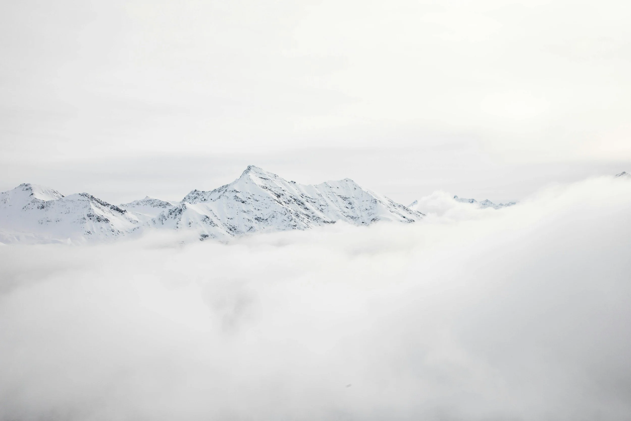 Montagnes enneigées au-dessus d'une couche de nuages ou de brouillard, sous un ciel gris clair.