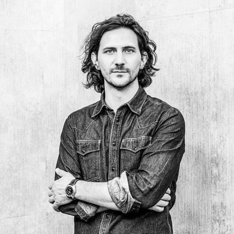 Serge Massat, associé Soqio Evaluation, portant une chemise en denim, se tenant contre un mur en arrière-plan, en noir et blanc.