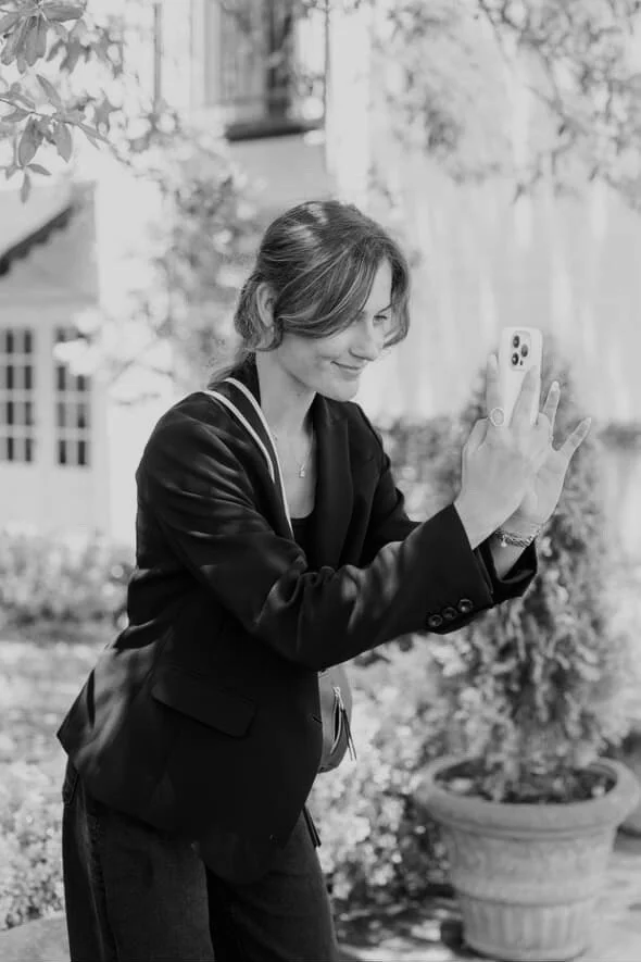 A woman with shoulder-length hair taking a selfie outdoors in a garden area.