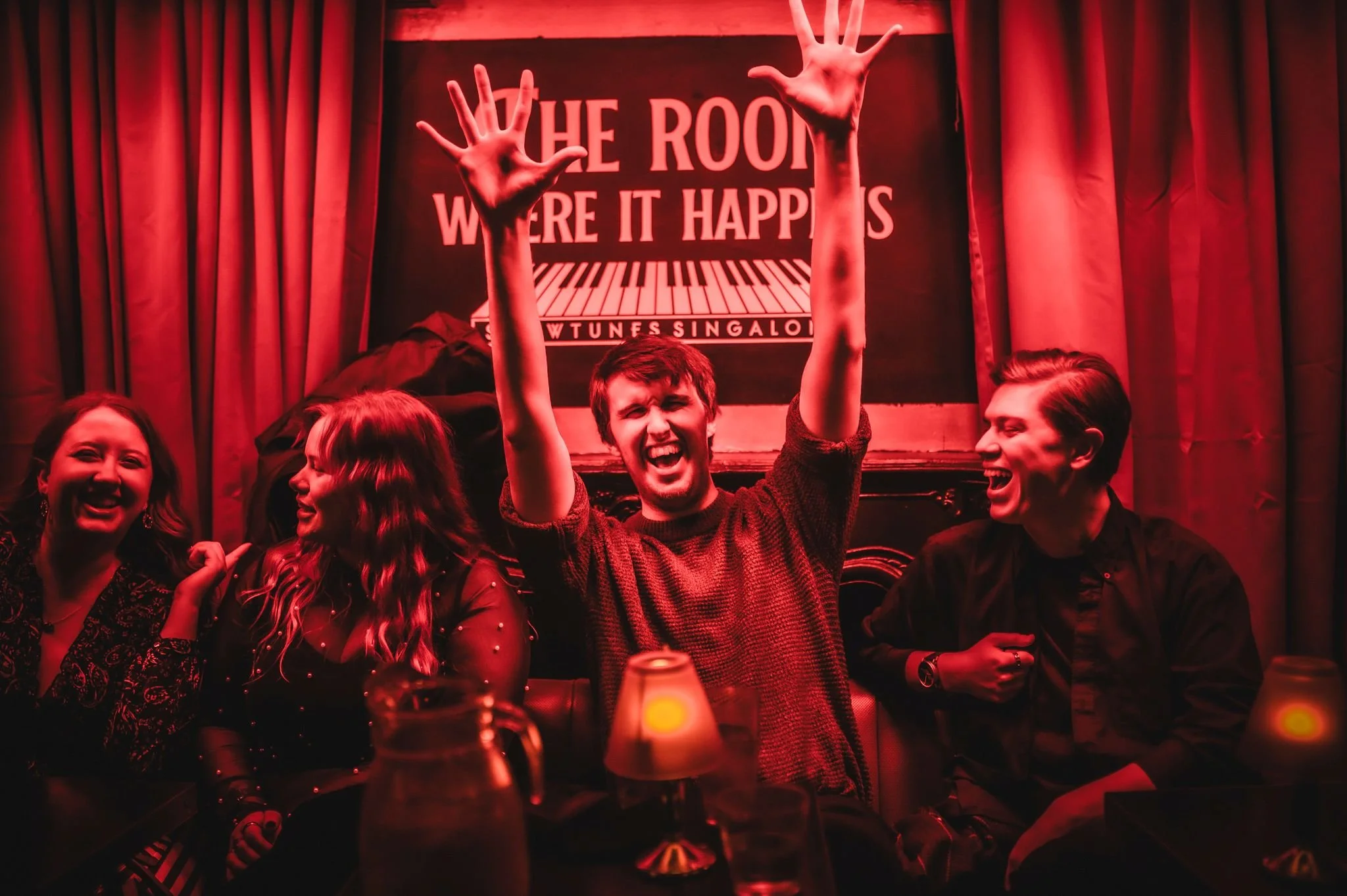 A group of four friends laughing and enjoying themselves at a bar or karaoke room, with red lighting and a sign in the background that reads 'The Room Where It Happens'.