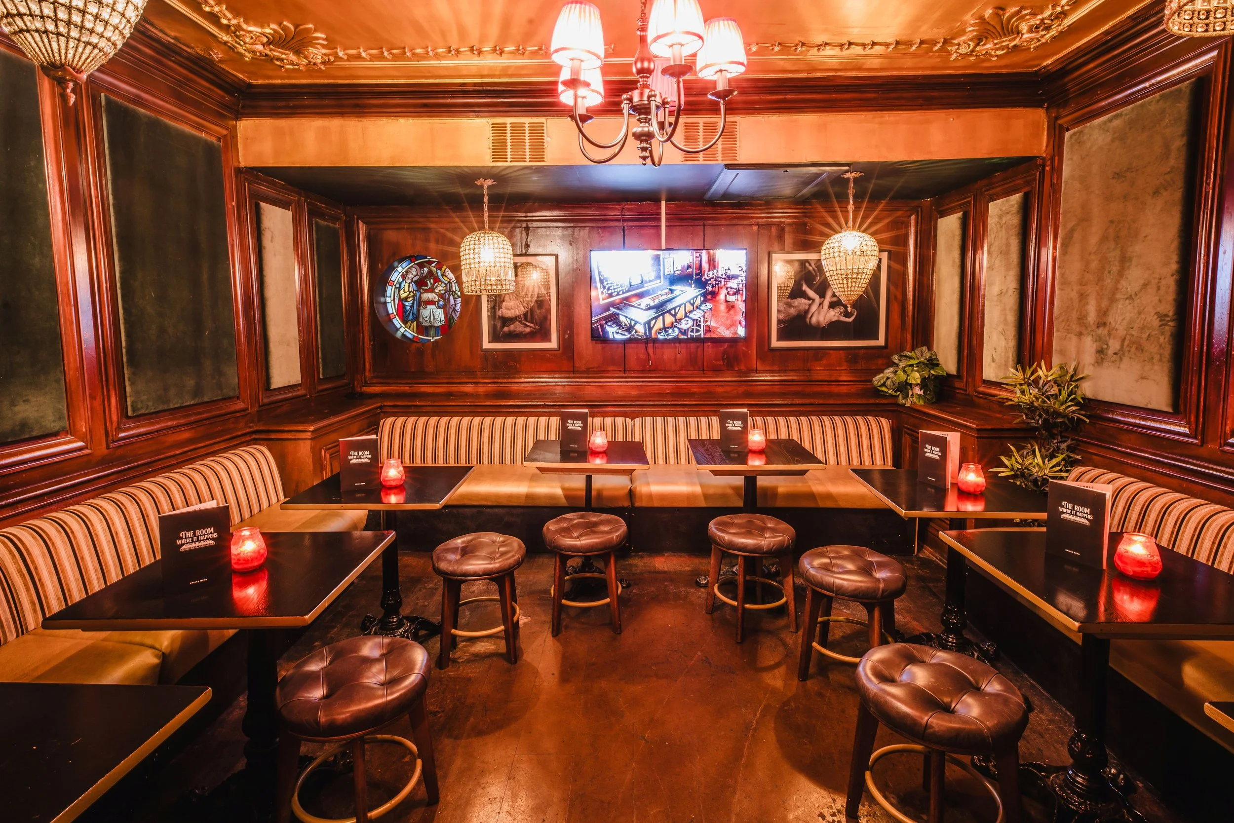 Cozy restaurant with dark wood paneling, striped upholstered benches, round leather stools, dim red lighting, and artwork on the walls including stained glass and photographs.