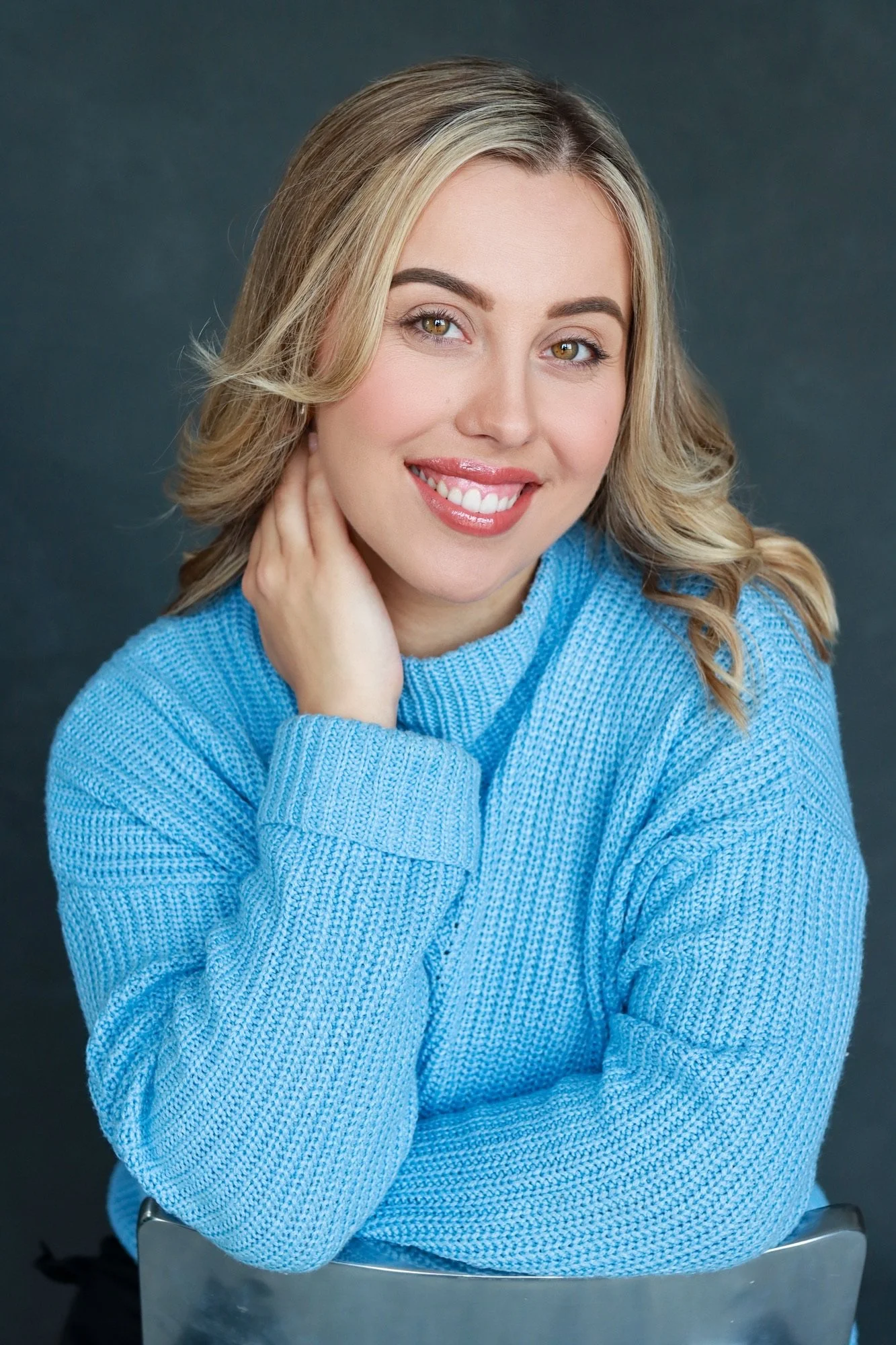 A woman with blonde hair, wearing a blue sweater, smiling and posing with her hand resting on her neck against a dark background.