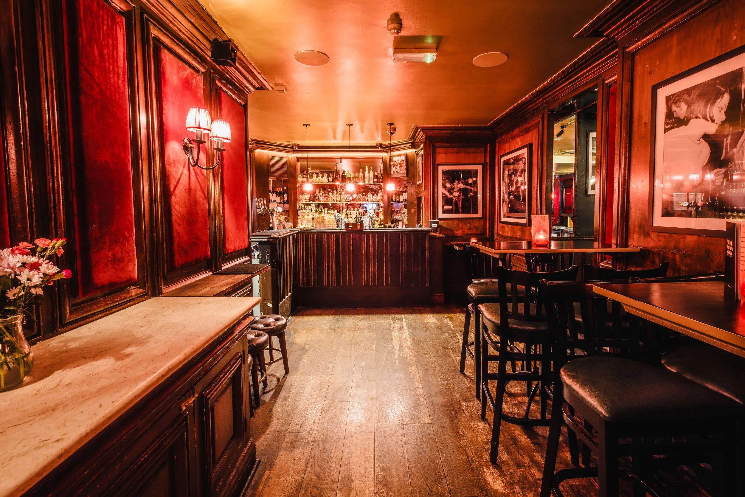 Interior of a cozy, dimly lit bar with wooden paneling, red velvet wall accents, framed black and white photos, and seating for patrons.
