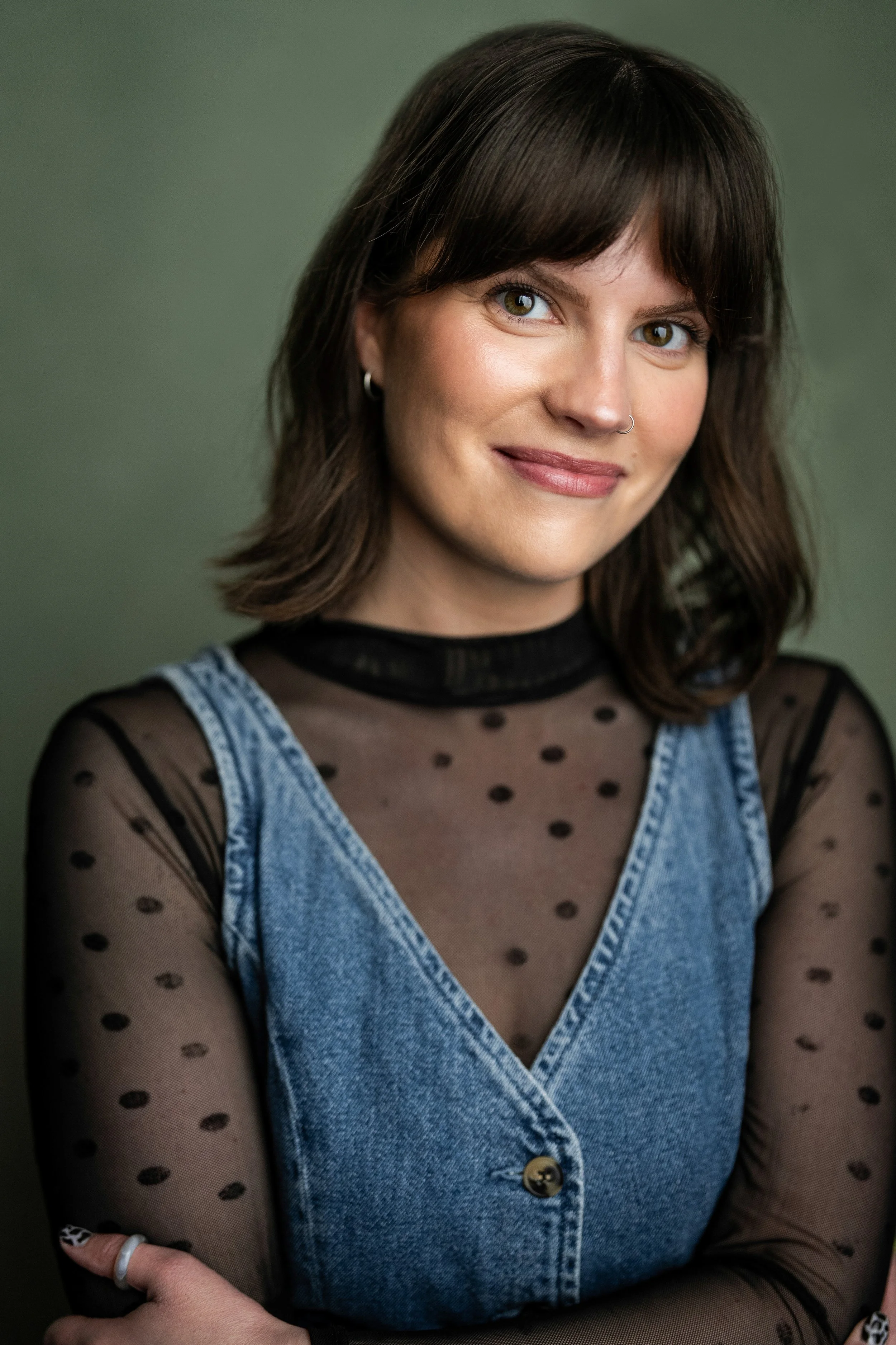 A woman with short brown hair, blue eyes, and a nose ring, smiling, wearing a black sheer polka dot top with a denim sleeveless jacket against a green background.