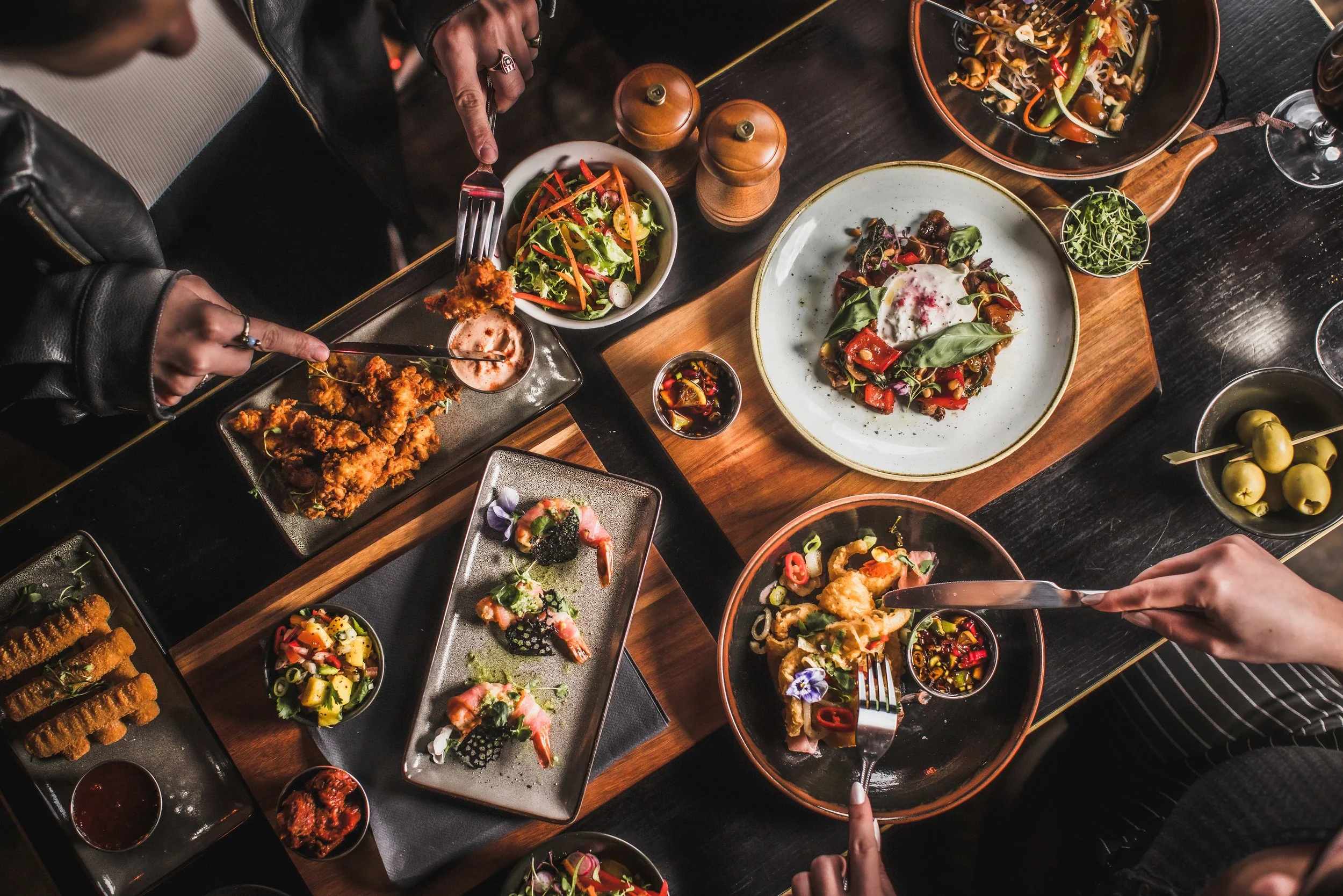A top-down view of a table with various dishes including salads, fried foods, and a small bowl of olives, with people serving themselves from the dishes.