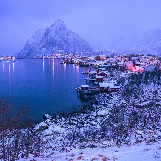 Snow-covered houses along a fjord with mountains in the background during twilight.