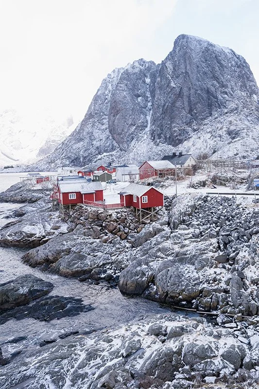 Snow-covered coastal village with red and white buildings, rocky shoreline, flowing water, and towering mountains in the background.