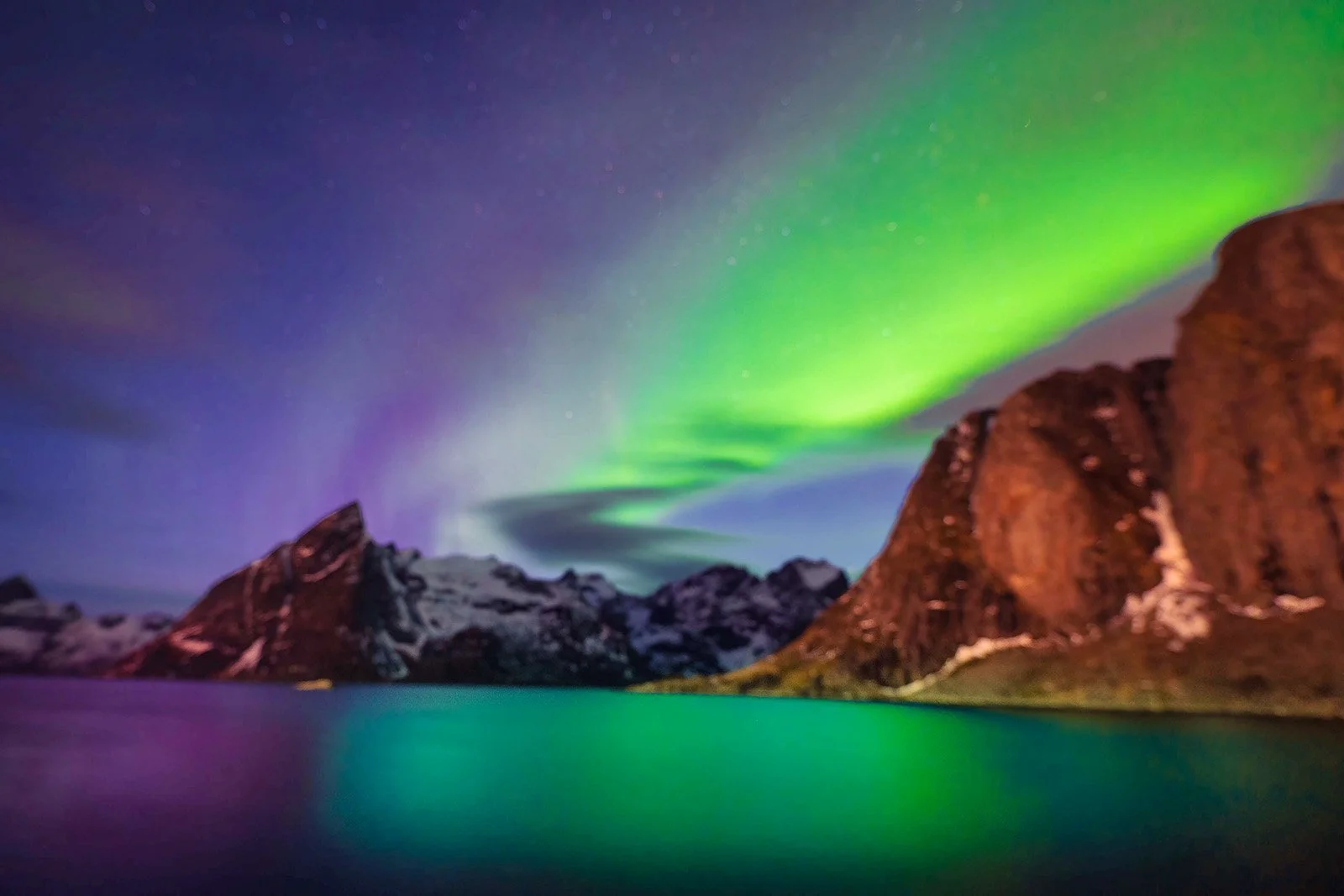 Northern Lights over a mountain lake with snow-capped peaks and rocky cliffs.