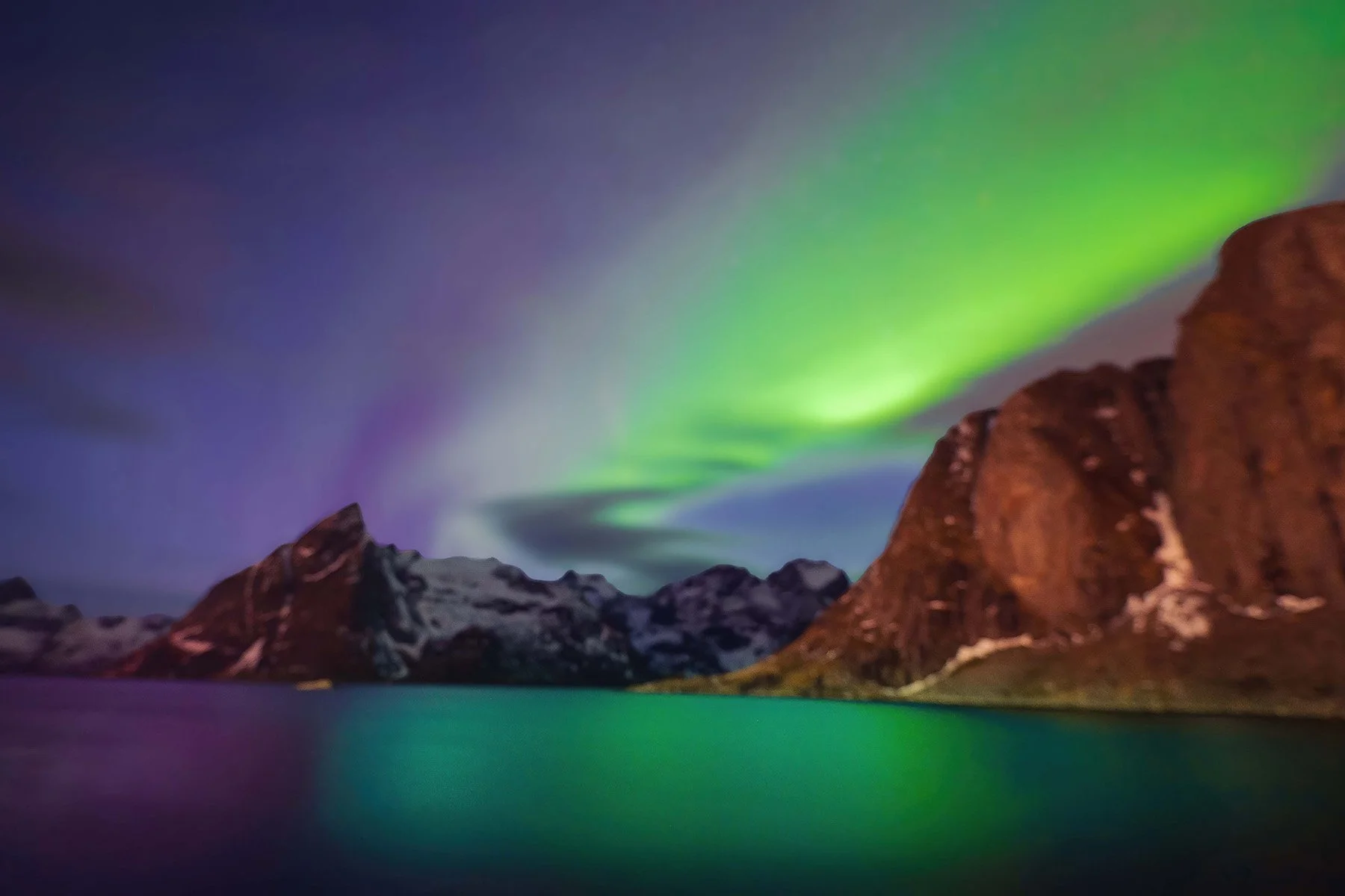 Northern lights over snow-capped mountains and a calm body of water at night.