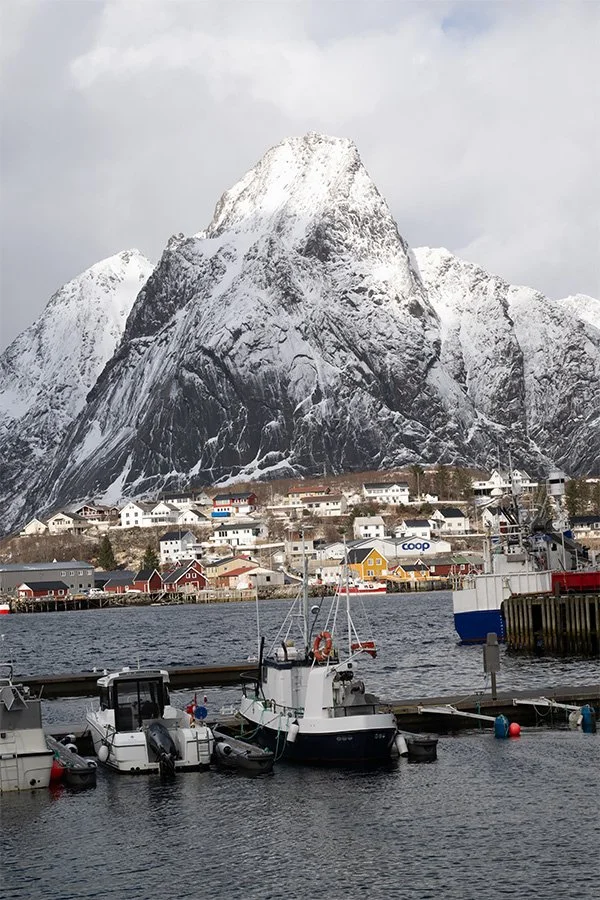A scenic view of a harbor with boats docked, a small coastal town with colorful houses, and a snow-covered mountain in the background, under a cloudy sky.