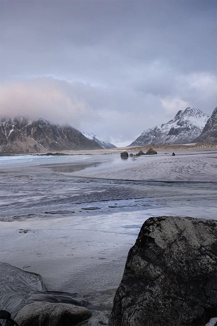 Snow-capped mountains and a cloudy sky reflecting on a partially frozen beach with rocks in the foreground.
