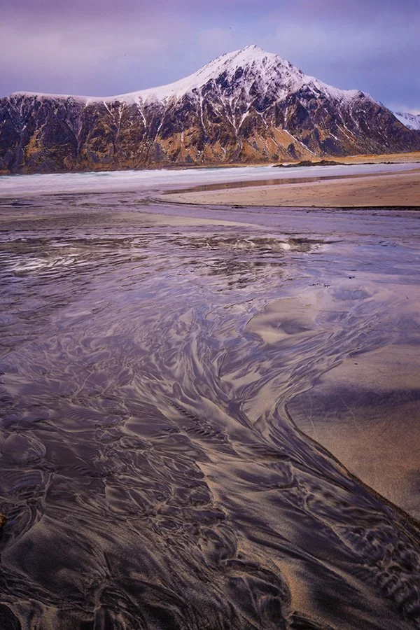 A snow-capped mountain behind a wide sandy beach with swirling water and patterns in the sand.