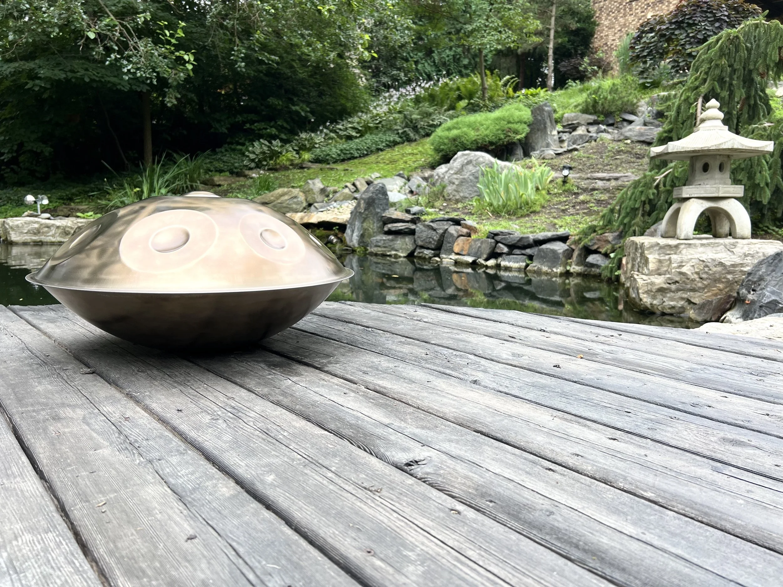 A metallic singing bowl on a weathered wooden deck overlooking a garden pond with rocks, greenery, and a small stone lantern in the background.
