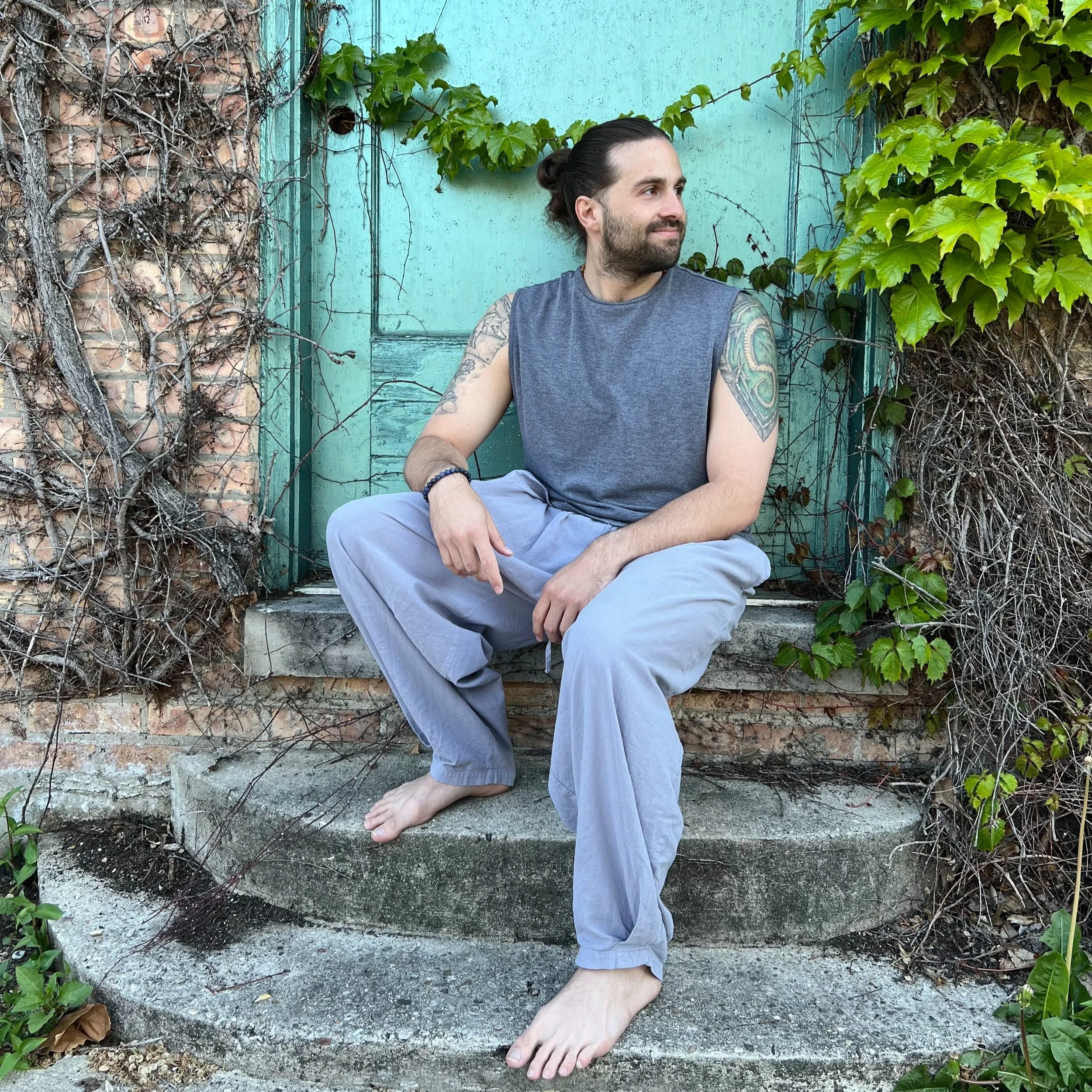 A man with long dark hair tied in a bun, tattooed arms, and light skin sitting barefoot on concrete steps outdoors, against a weathered turquoise door and brick wall surrounded by ivy and green leaves.