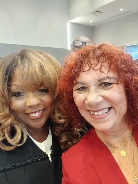 Two women smiling for a selfie, one with curly red hair and the other with blonde hair, in an indoor setting.