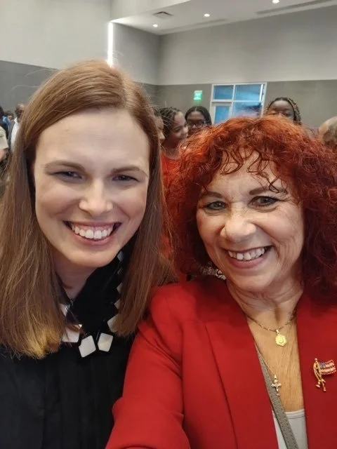 Two women smiling for a photo at an indoor event, with a crowd in the background.