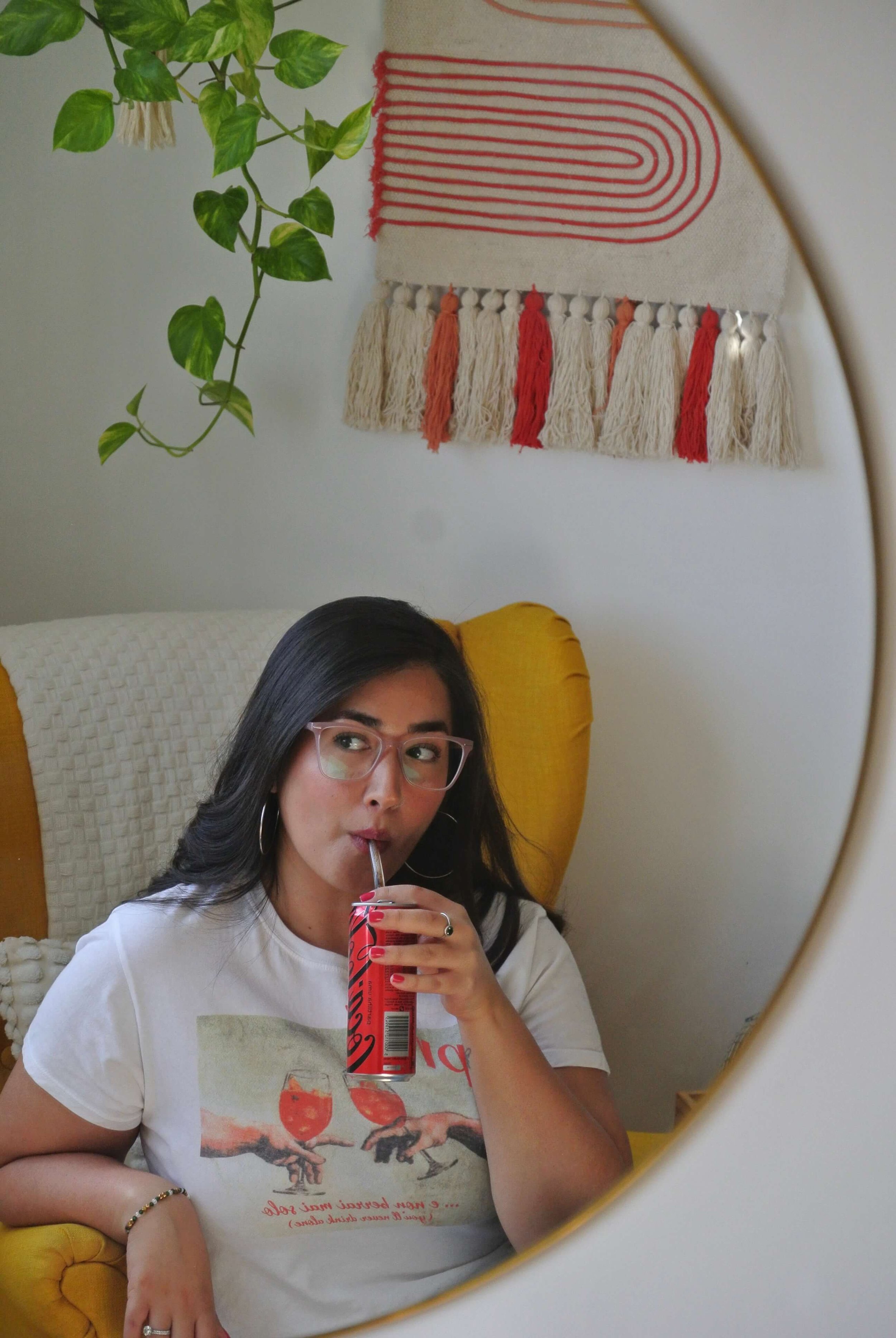 Ariel Delamora sitting in a yellow chair, sipping a drink through a straw and looking to the side in a cozy, creative home setting.