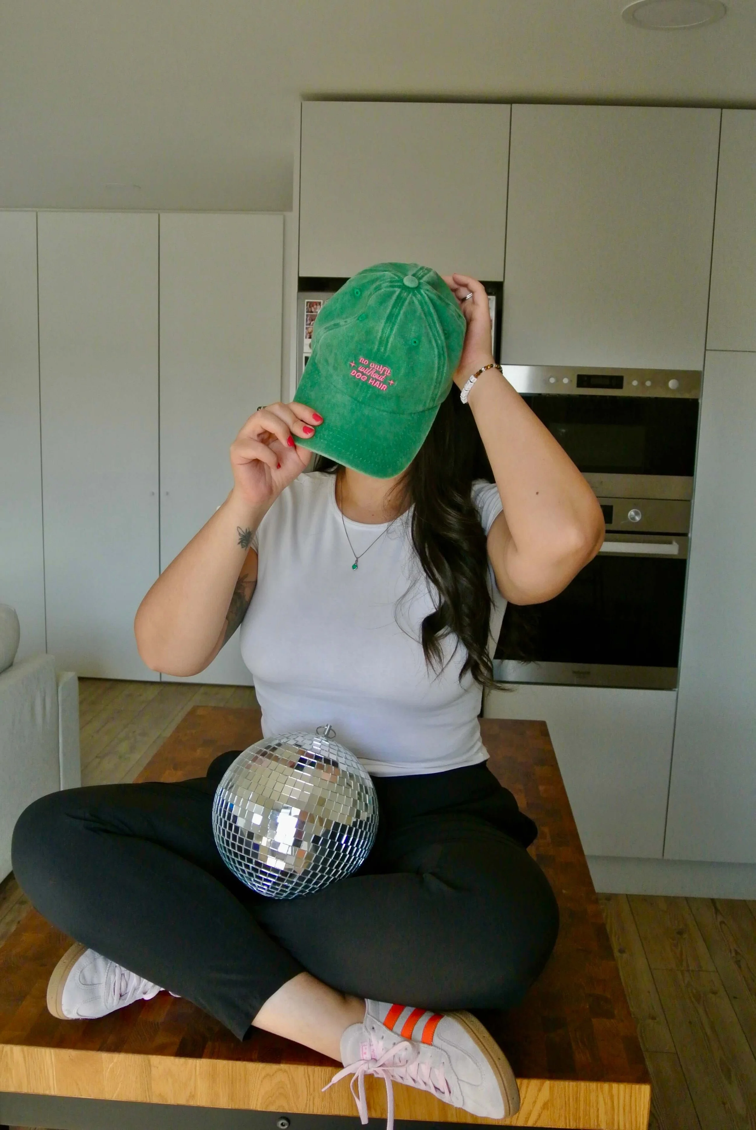 Ariel Delamora sitting cross-legged on a table holding a disco ball and adjusting a green hat in a bright, modern kitchen setting.