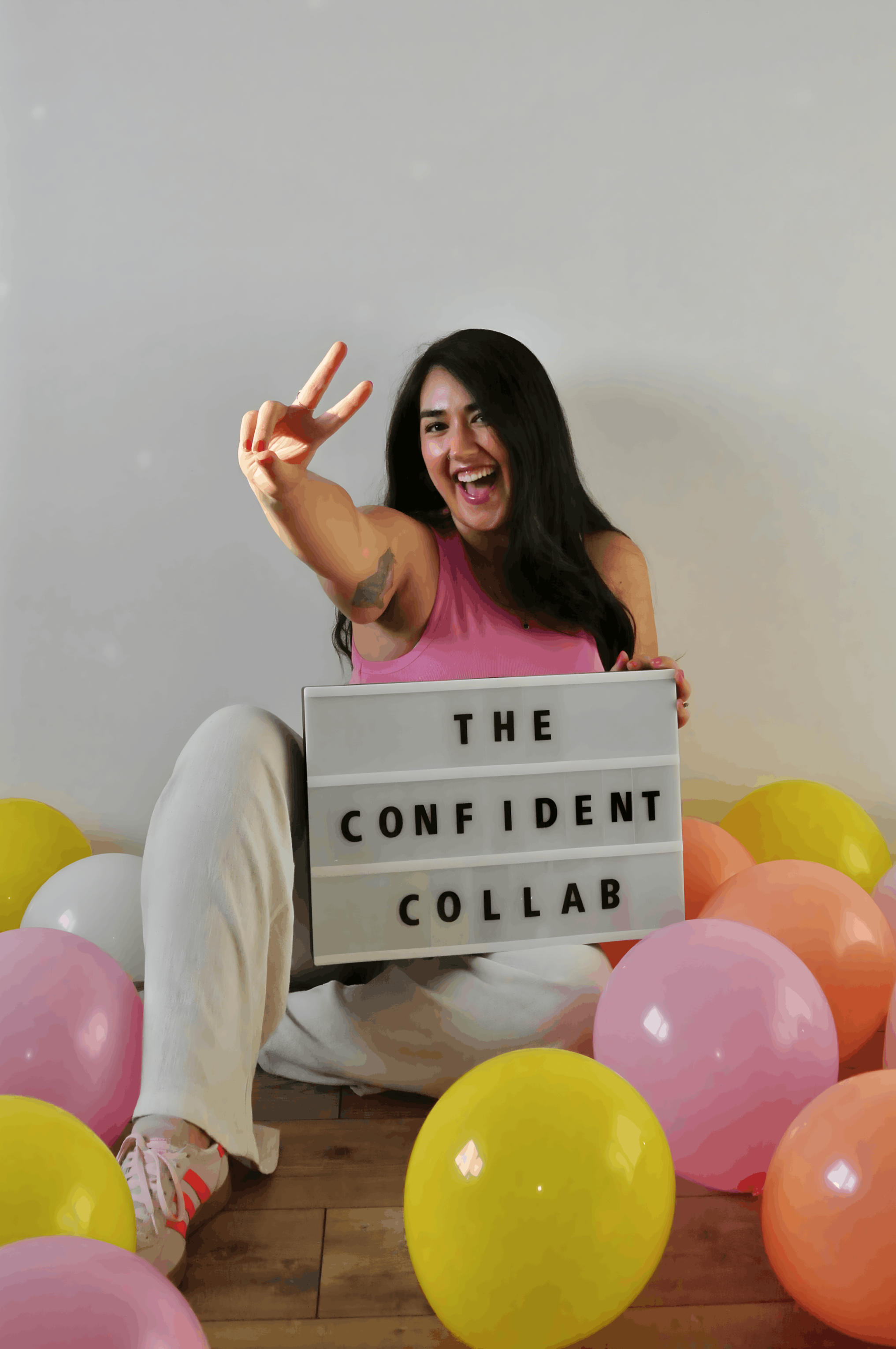 Ariel Delamora sitting among colorful balloons holding a sign that reads “The Confident Collab” and smiling while flashing a peace sign.