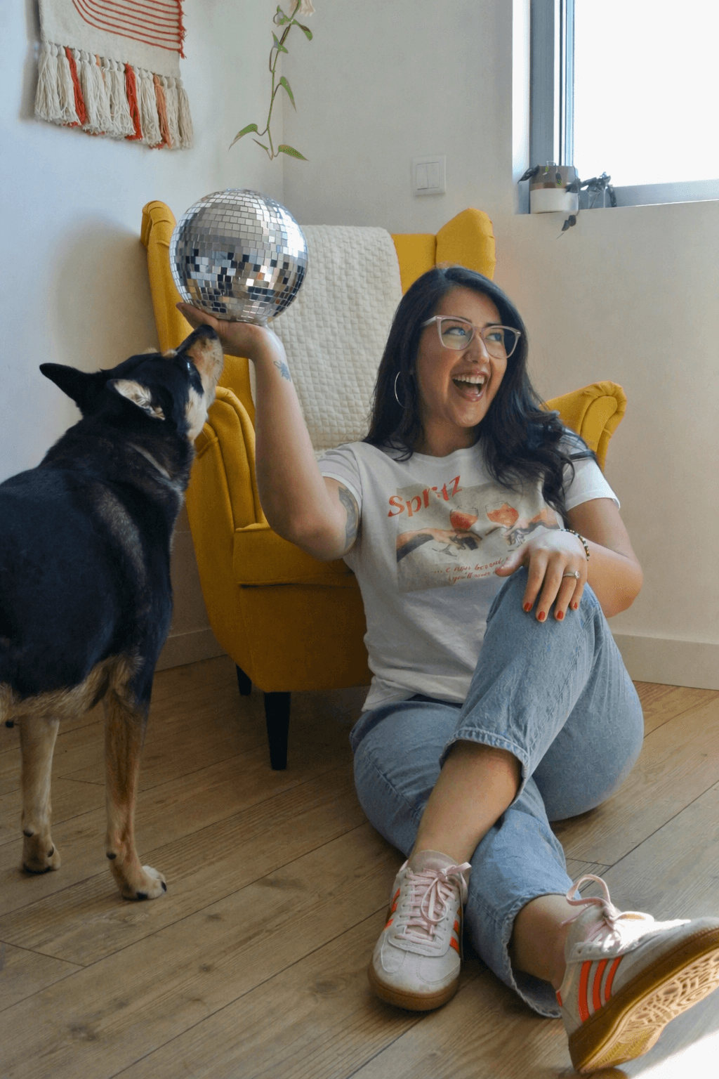 Ariel De La Mora sitting on the floor laughing while holding a disco ball, with her dog beside her in a cozy home setting.