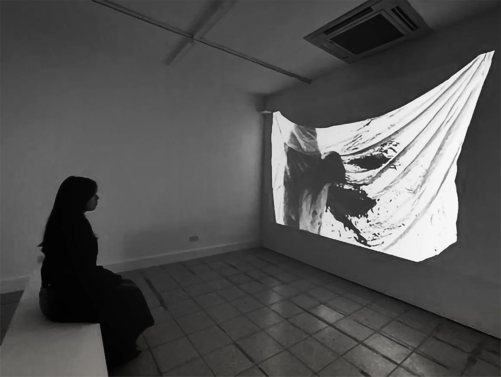 A woman sitting on a bench watching a black-and-white abstract projection on a wall in a minimalistic room.