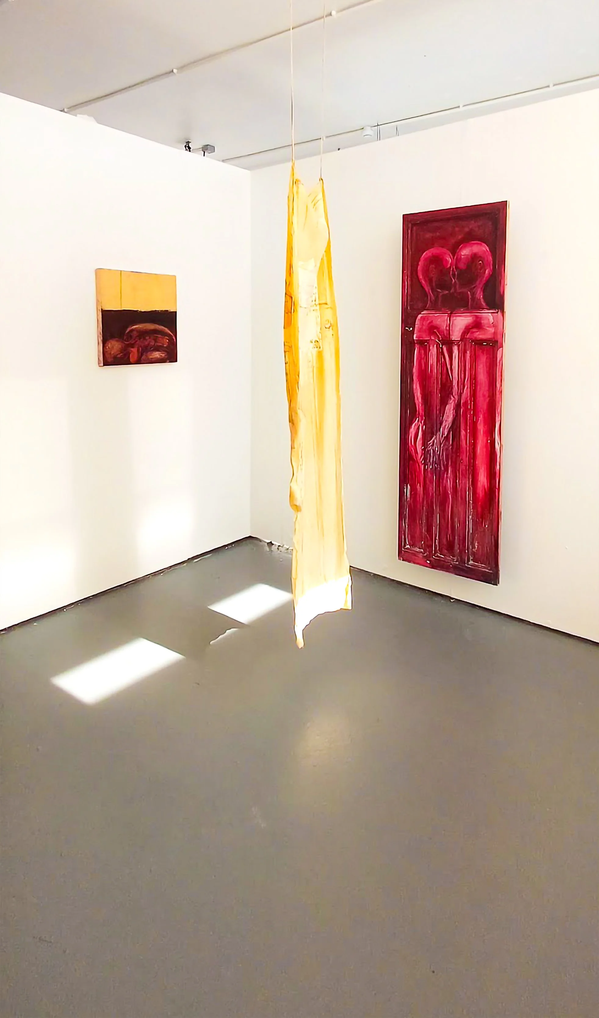 Art gallery with three paintings and a hanging yellow fabric or paper sculpture, illuminated by natural sunlight casting shadows on the gray floor.