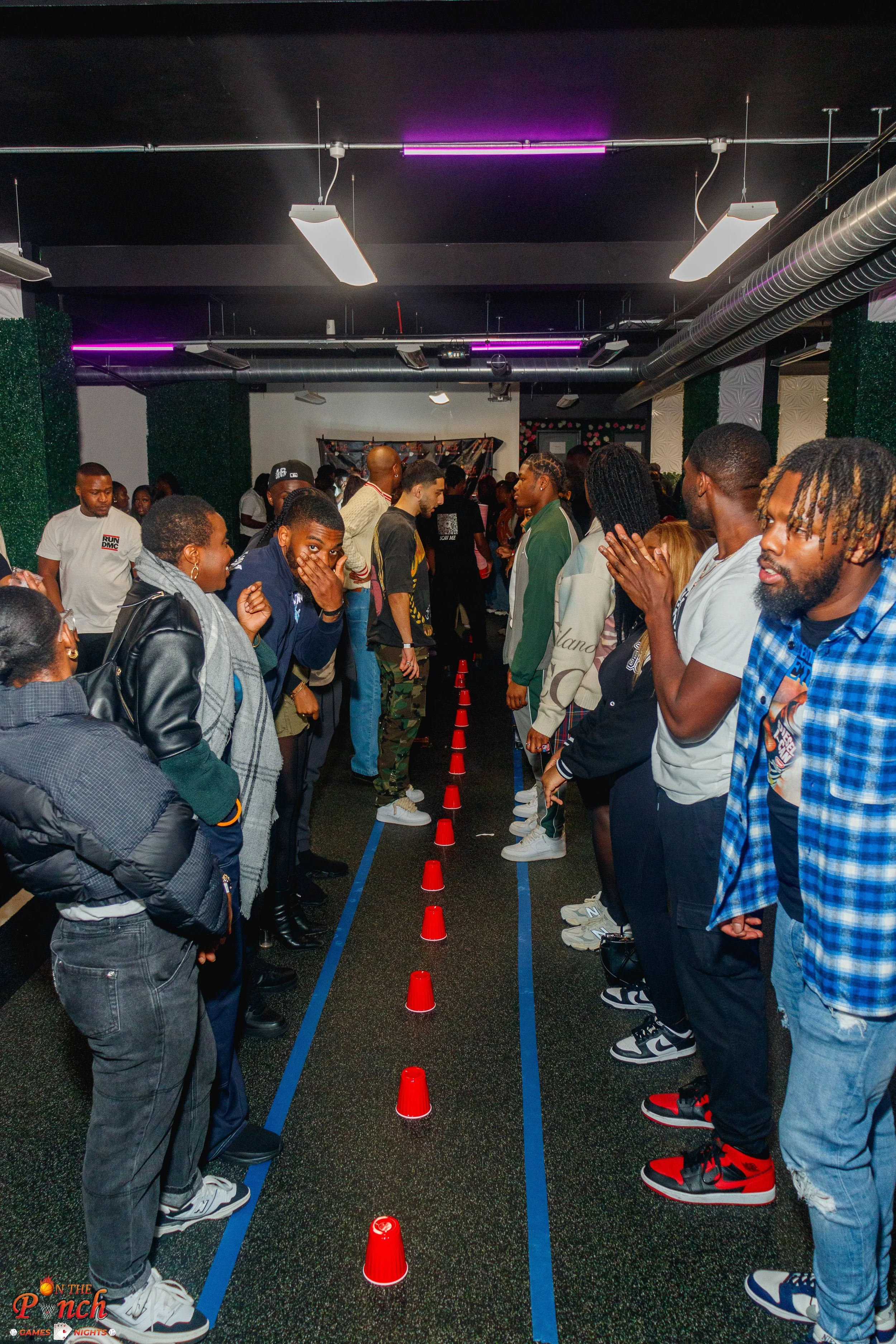 A group of people standing in two lines facing each other, separated by a row of red cones, possibly participating in a team-building activity or game indoors with overhead lighting and green walls.