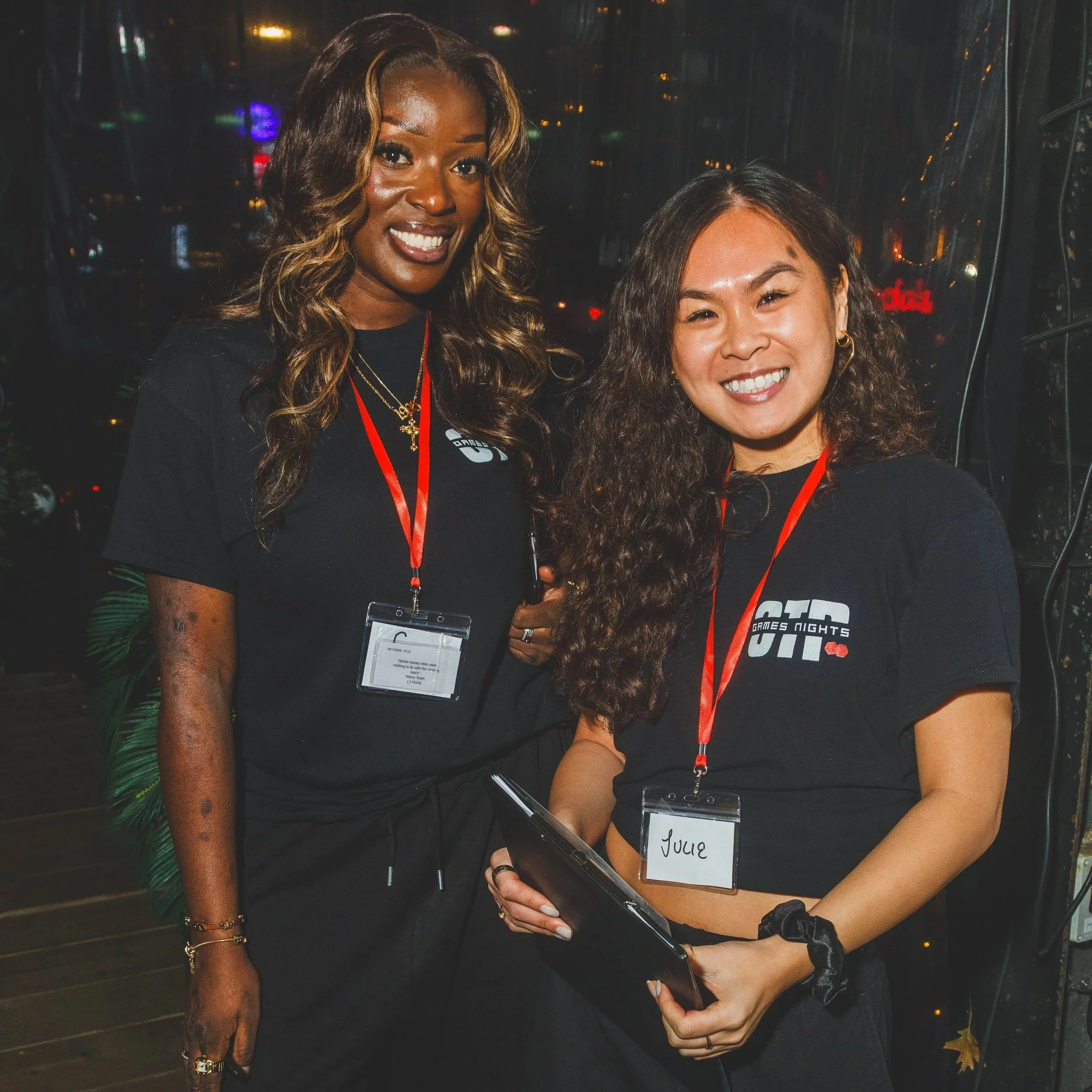 Two women standing together at an event, both wearing black T-shirts with 'GAMES NIGHTS' printed on them, and each wearing a red lanyard with a name tag. The woman on the left has long curly hair and is smiling, while the woman on the right has long wavy hair and is holding a tablet, also smiling.
