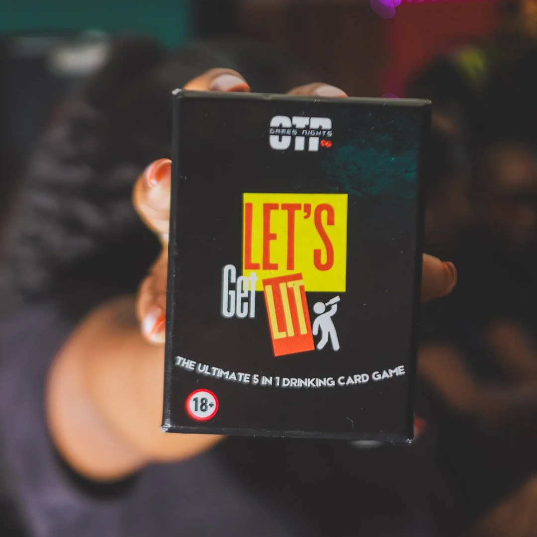 A group of seven friends gathered at a table playing a social game outdoors, with string lights overhead. They are smiling and laughing, with drinks in front of them, and a man in the center holding a card that says, "Never Lny I Ever...". The game cards on the table include "Do It Or Drink!", "Ice Breakers", "Lucky Dip", "Trivia", and "Never!".