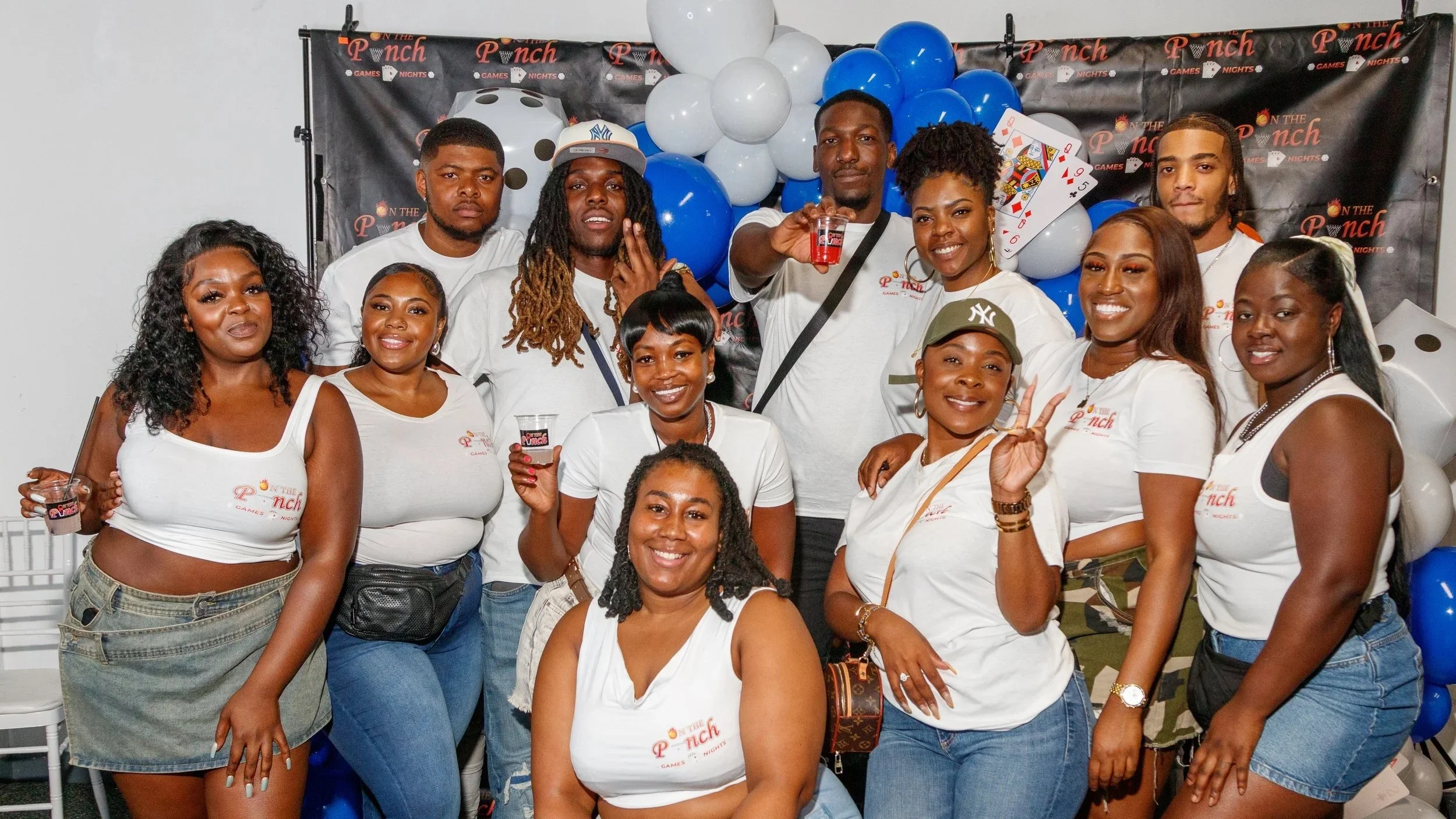 A group of people in white shirts celebrating at an event with a black and red backdrop that reads 'On the Pnch,' surrounded by blue and white balloons, holding drinks, and posing for a photo.