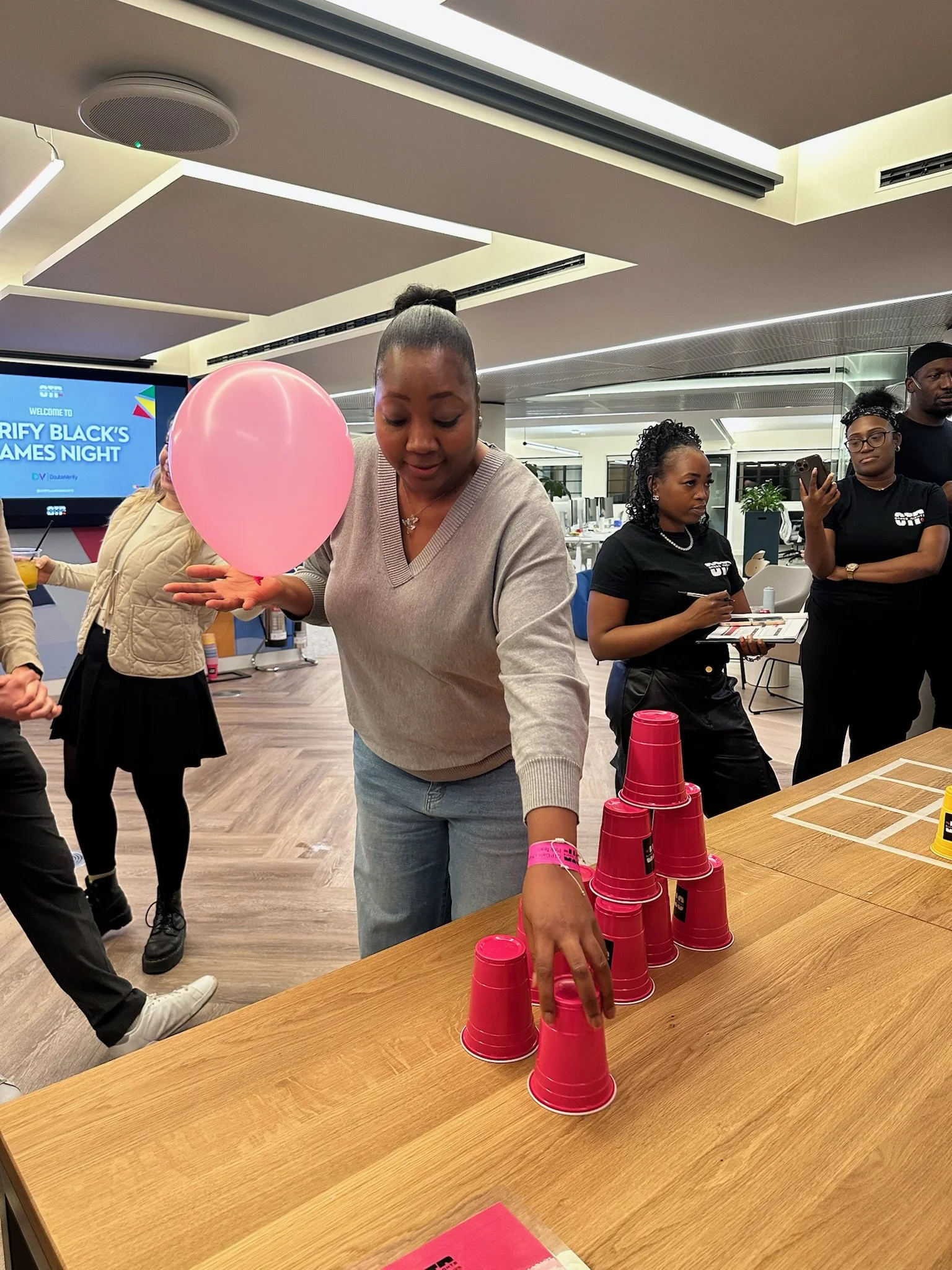 A woman stacking pink plastic cups on a wooden table during a social event in an indoor setting.