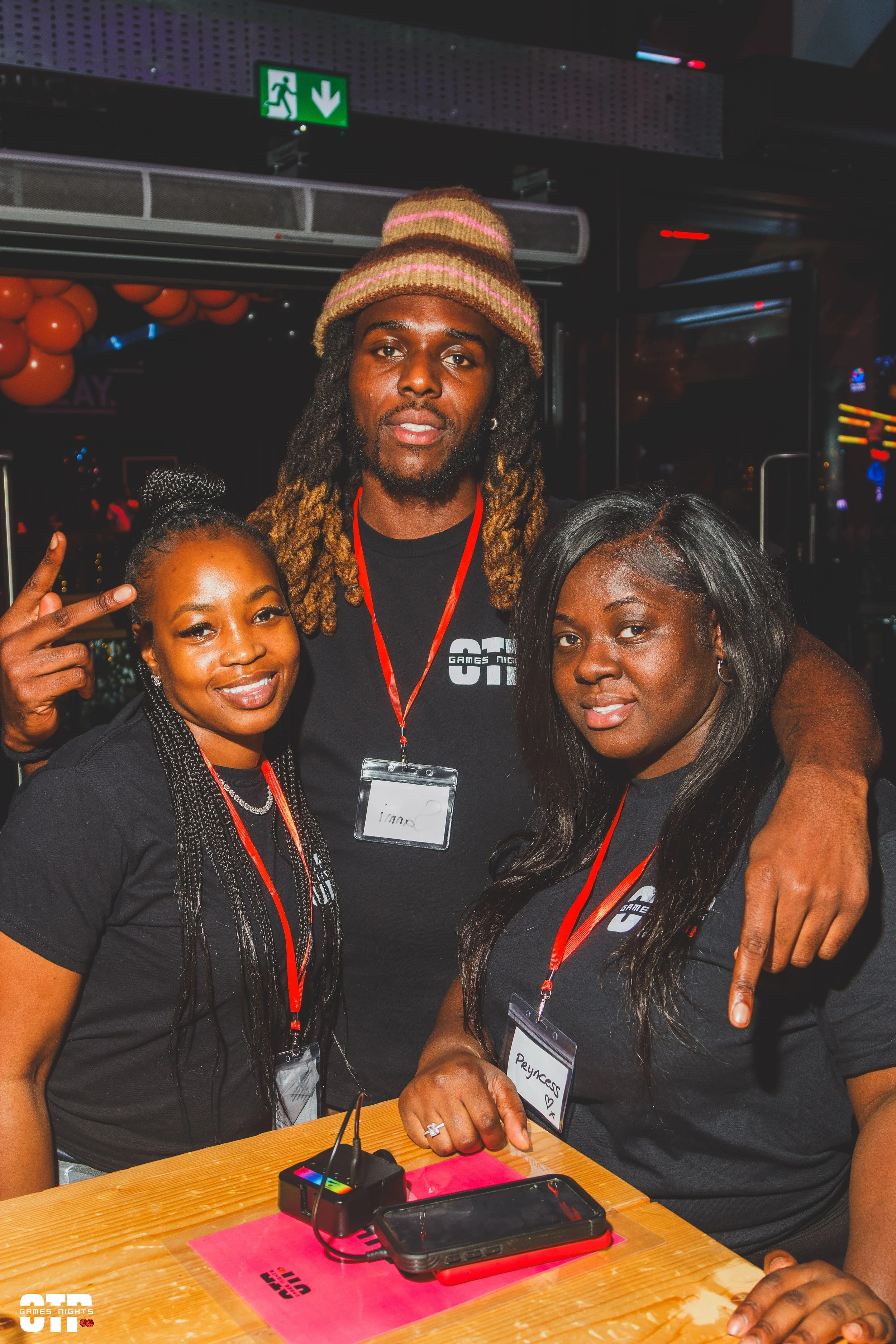Three people at an event, wearing black shirts with a logo, standing close together and smiling; one man in the center with dreadlocks and a hat, flanked by two women with long hair, one with braided hair and the other with straight hair. They are indoors, with a wooden table in front of them and an exit sign above.
