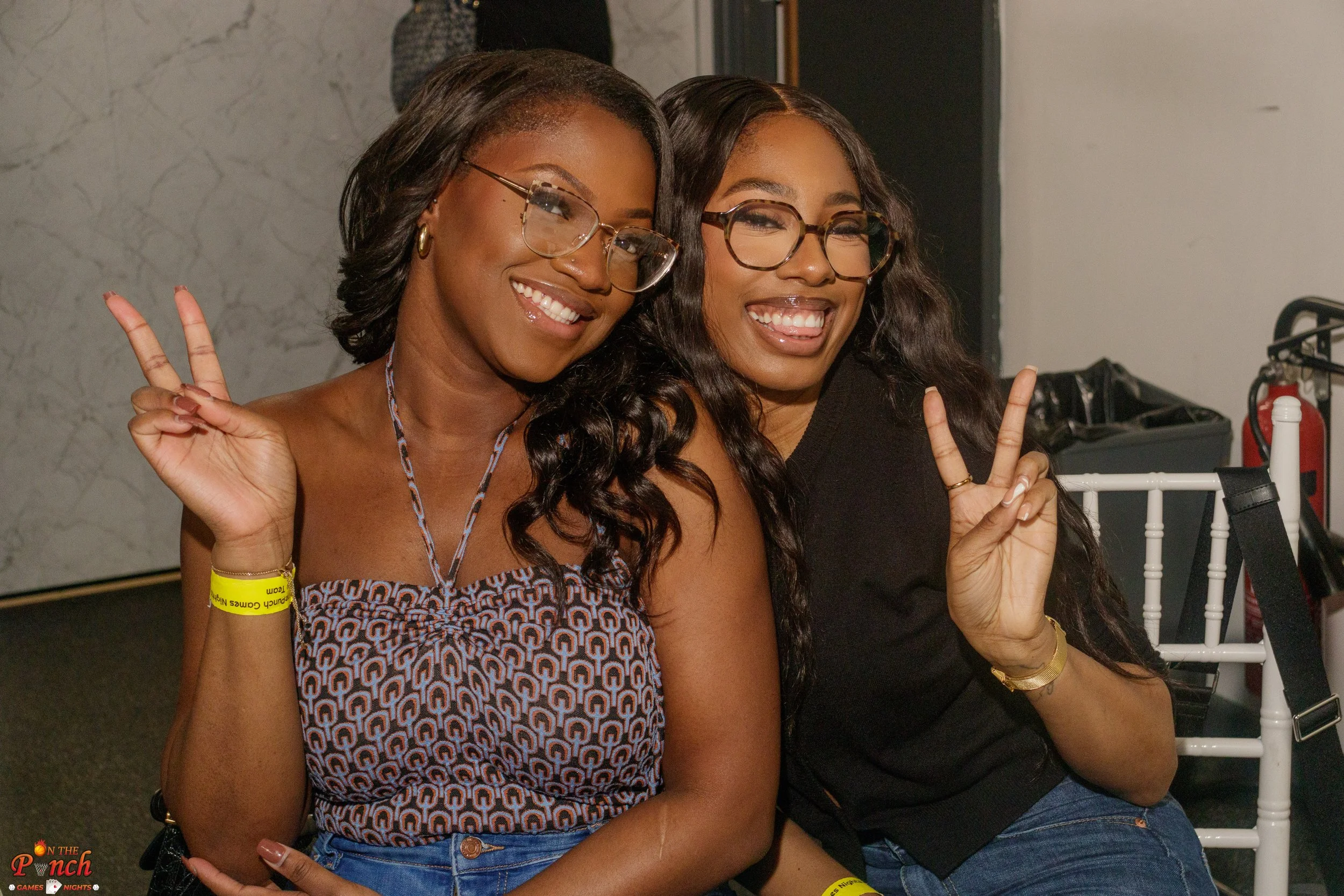 Two women sitting side by side smiling and making peace signs with their hands. Both wearing glasses, one in a patterned dress and the other in a black shirt.