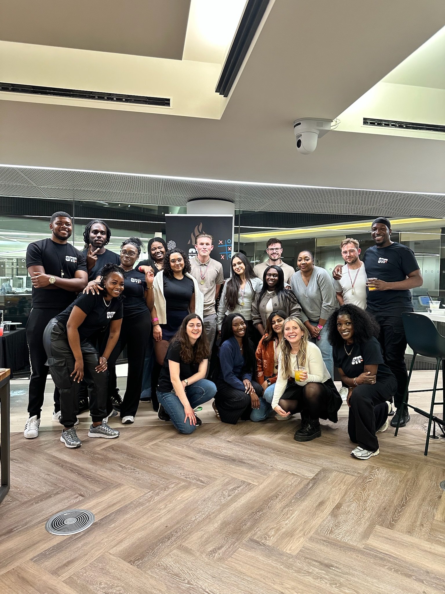 Group of six people celebrating, wearing medals, standing in front of a balloon backdrop with black, white, gold, and silver balloons.