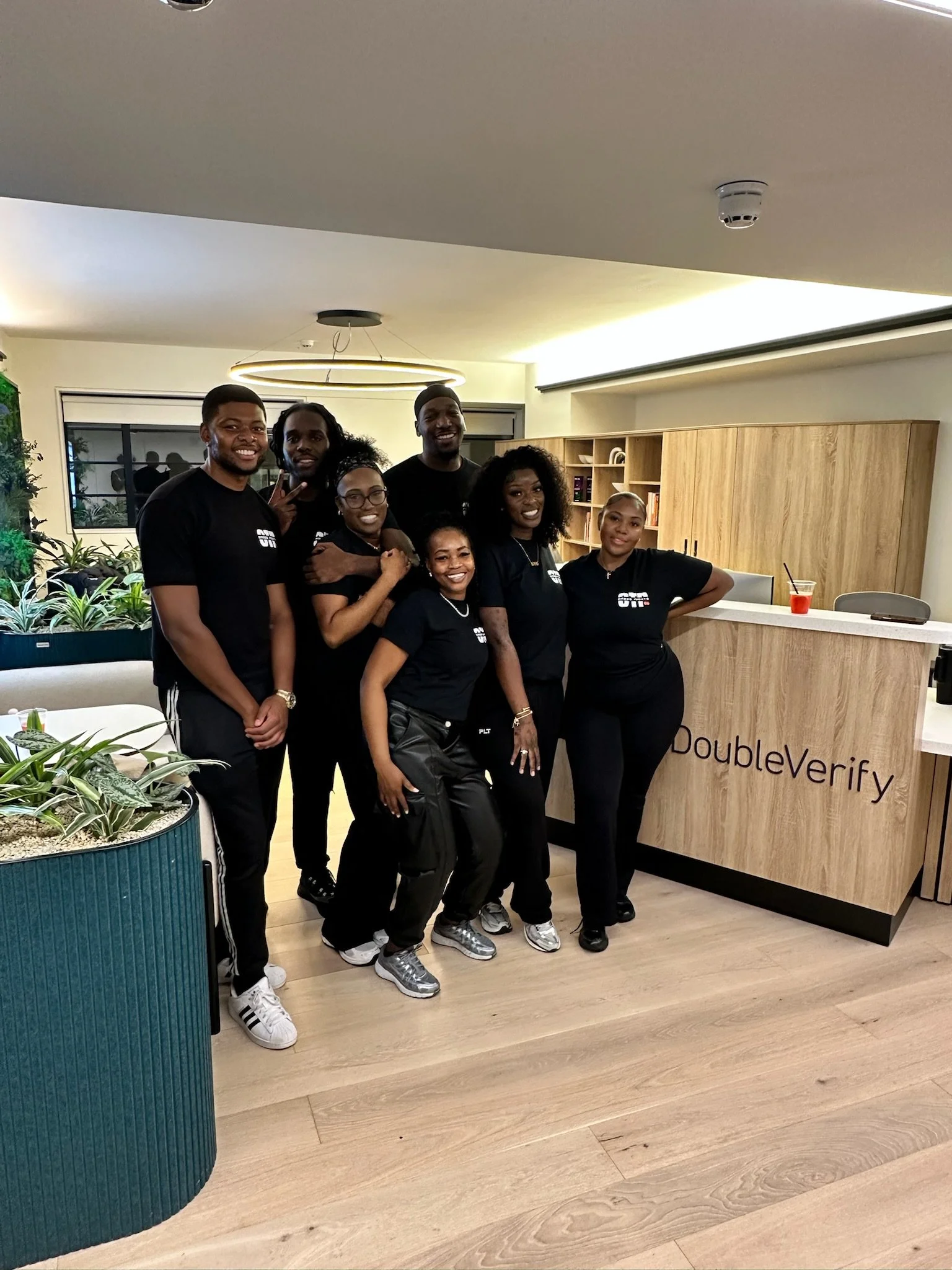 Group of people standing together in a lobby, smiling, with a reception desk that has the word 'DoubleVerify' on it. There are plants and a window in the background.