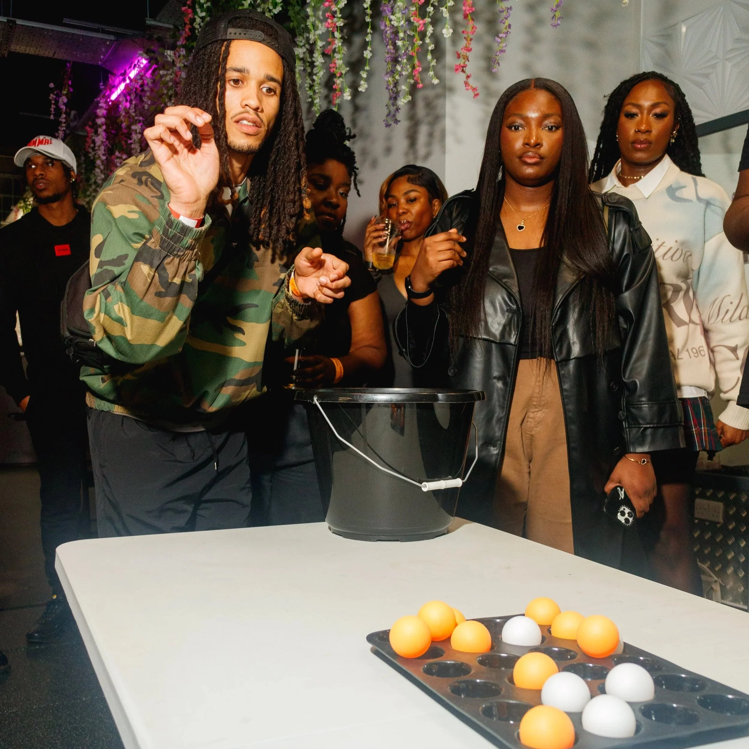 Group of people gathered around a table playing a game with eggs, some people are holding drinks, with colorful hanging flowers in the background.