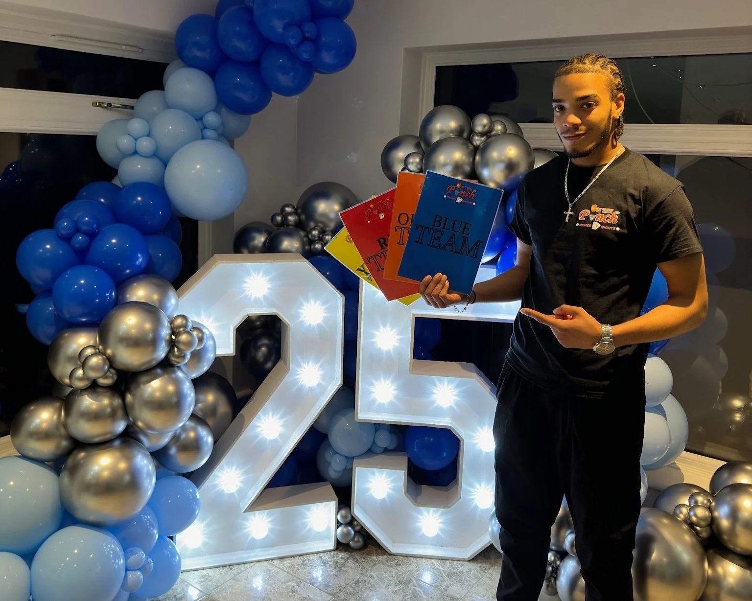 A young man holding colorful cards standing in front of a large illuminated number 21 surrounded by blue, silver, and white balloons at a celebration.