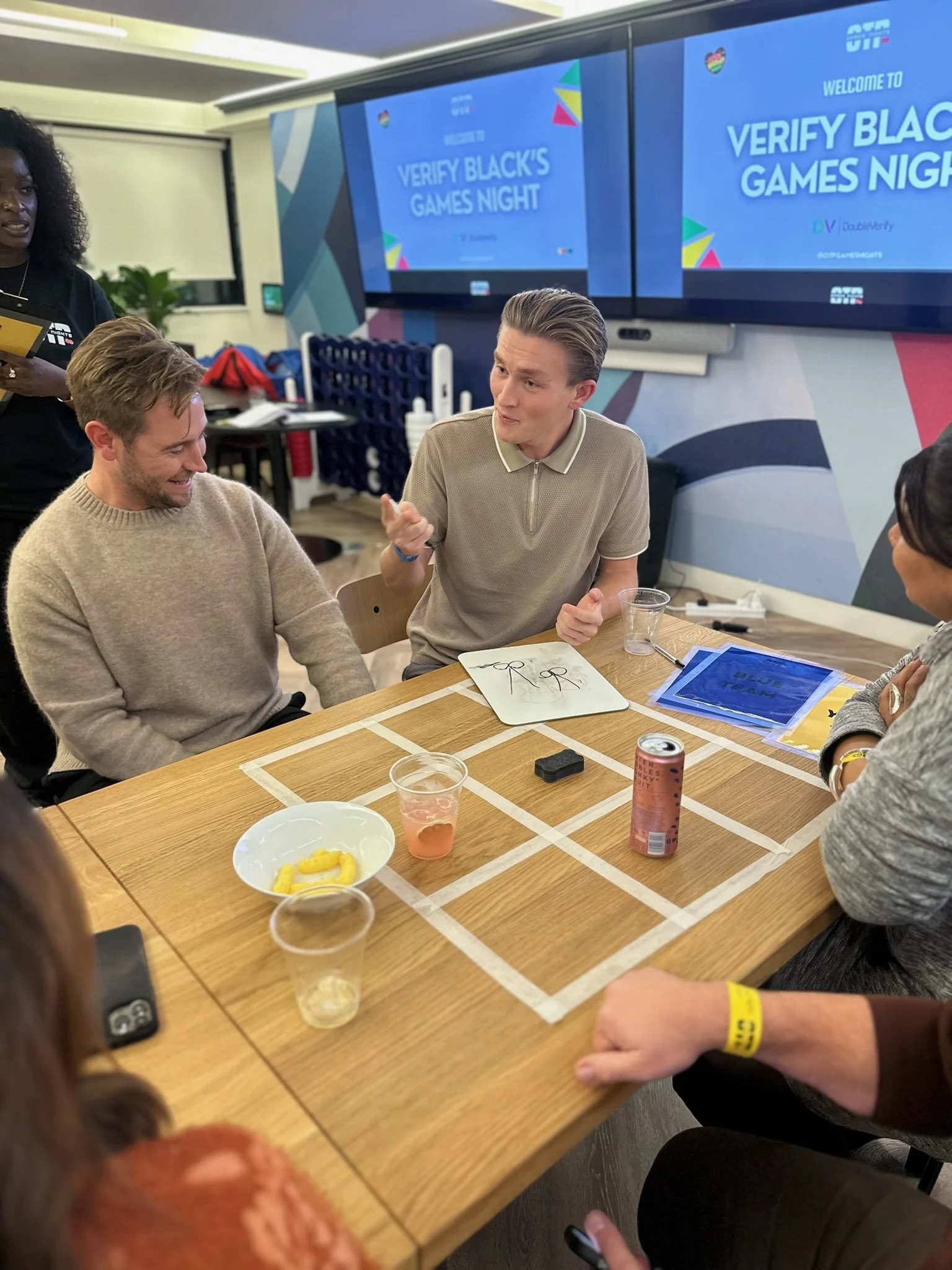 People sitting around a table playing a game during a social event called 'Verify Black's Games Night' with a large screen in the background displaying the same event name.